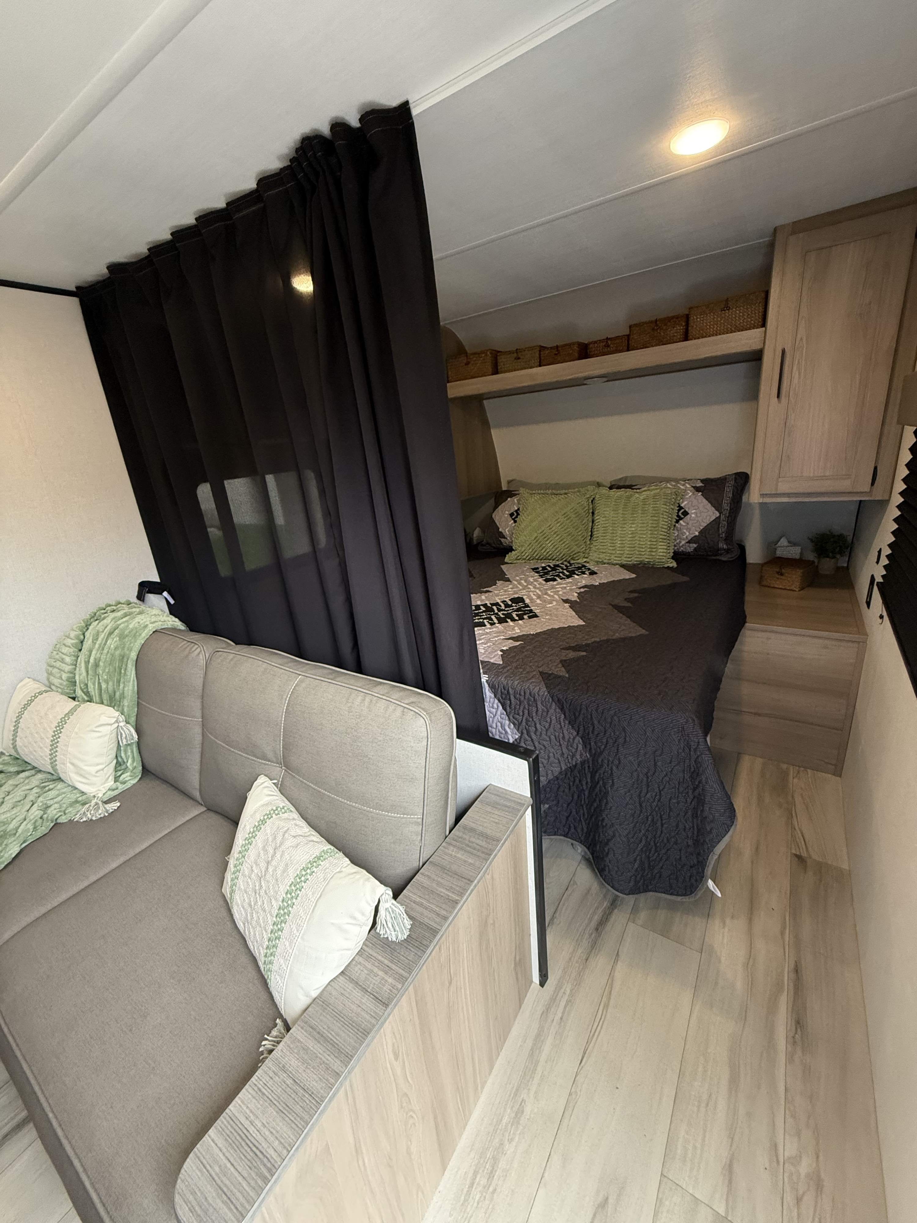 Cozy RV rental interior featuring a gray sofa, privacy curtain, and a comfortable bed with overhead storage and lighting.. Keystone RV 23B 2025