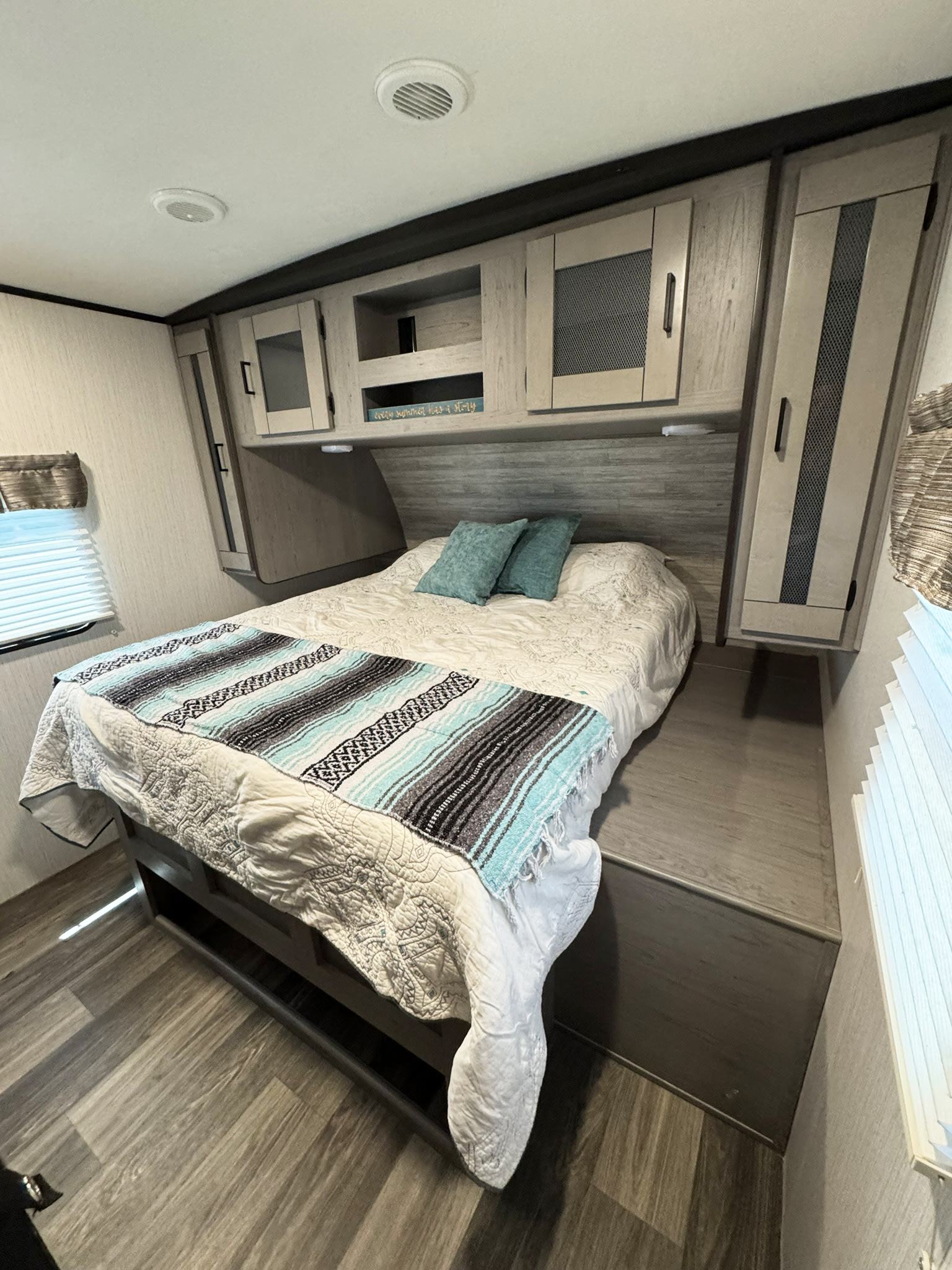 Modern RV rental bedroom featuring a bed with a striped blanket, overhead cabinets, window, and wood-look flooring.. Heartland RVs Mallard 2021