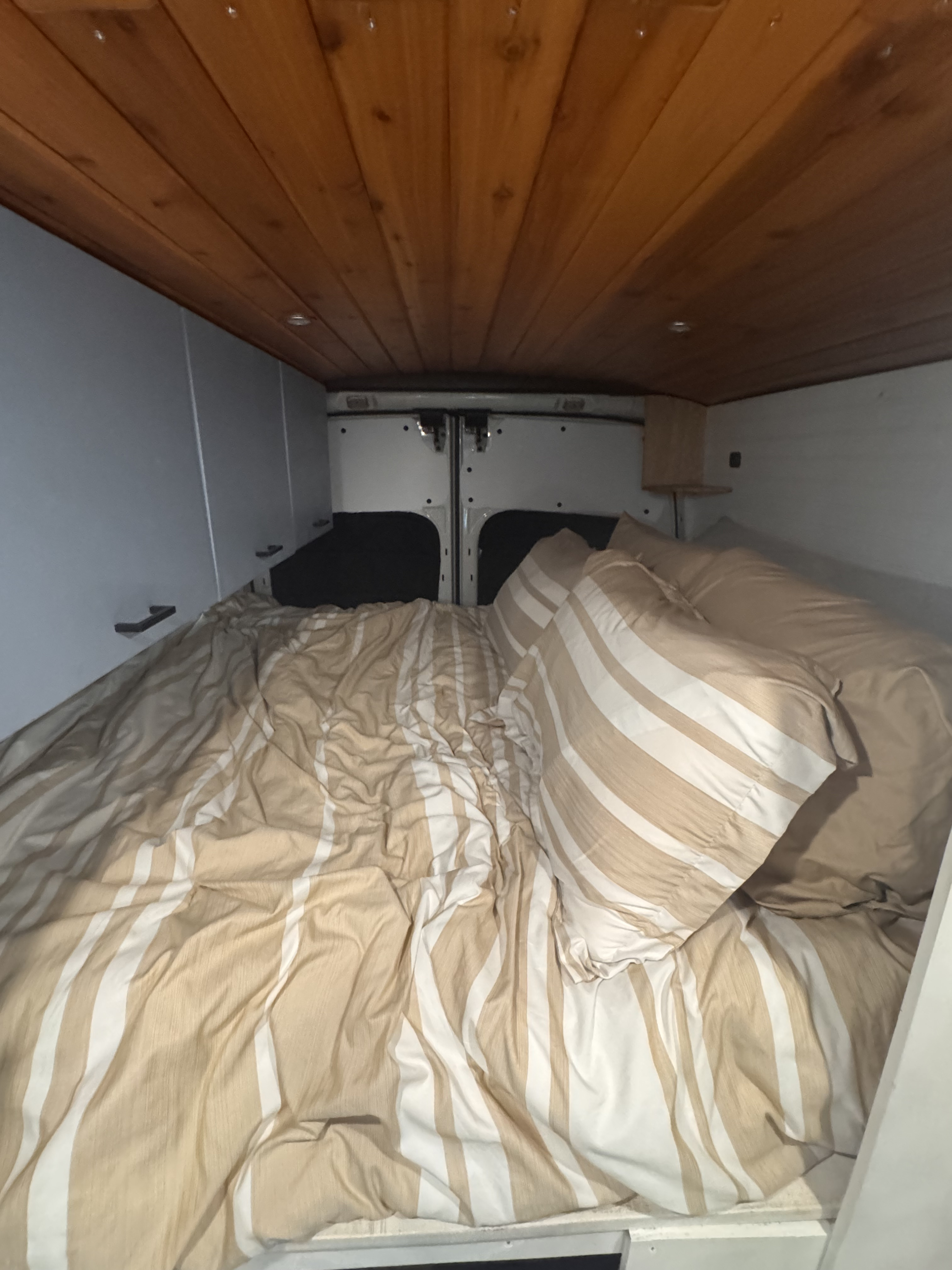 Interior of an RV rental: a cozy bed with striped bedding, wood ceiling, and storage cabinets.. Ford Van 2015
