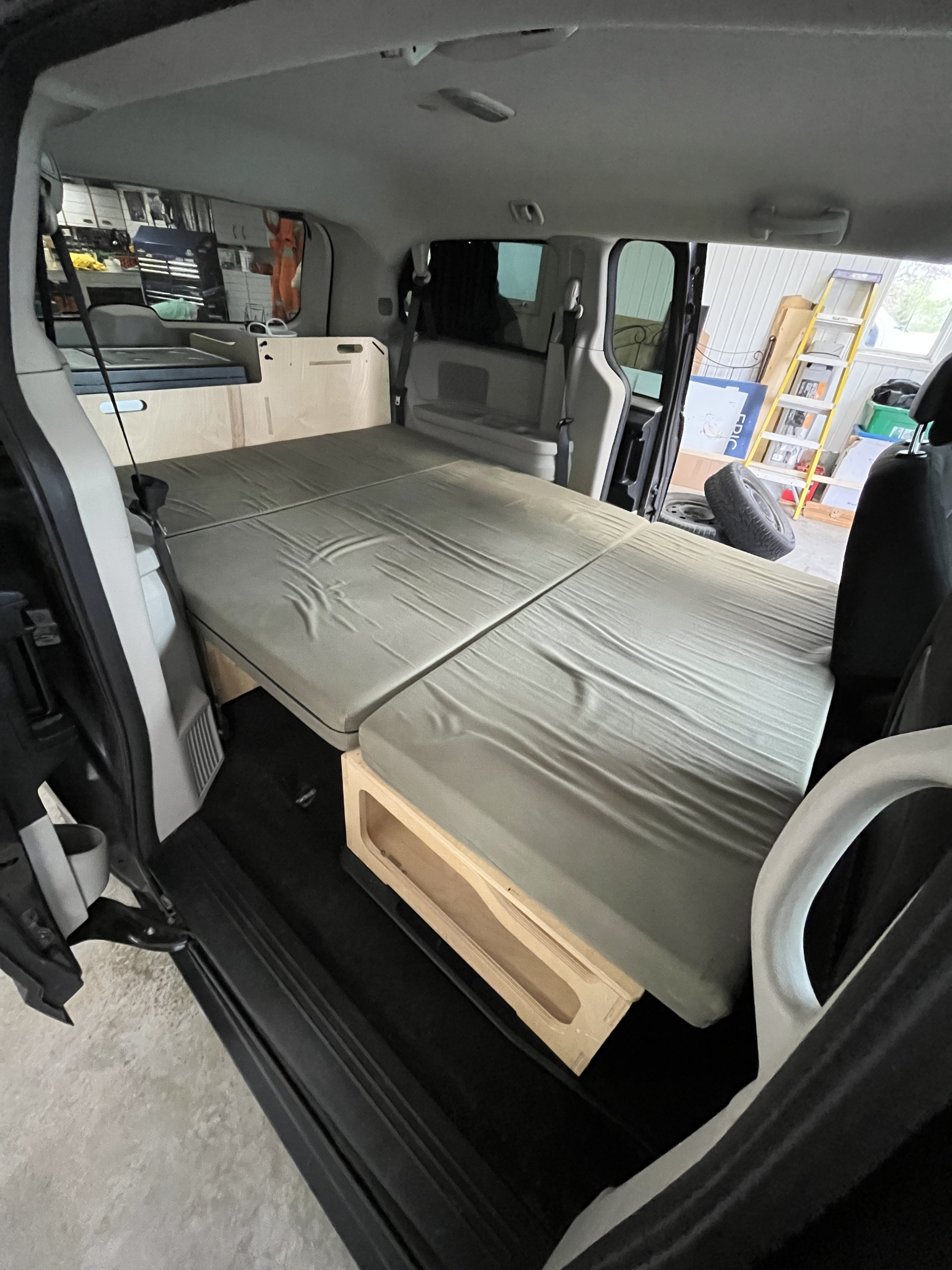 View inside a camper minivan, showing a folded gray mattress on a custom wooden bed platform, ideal for an RV rental.. Dodge Grand Caravan 2015