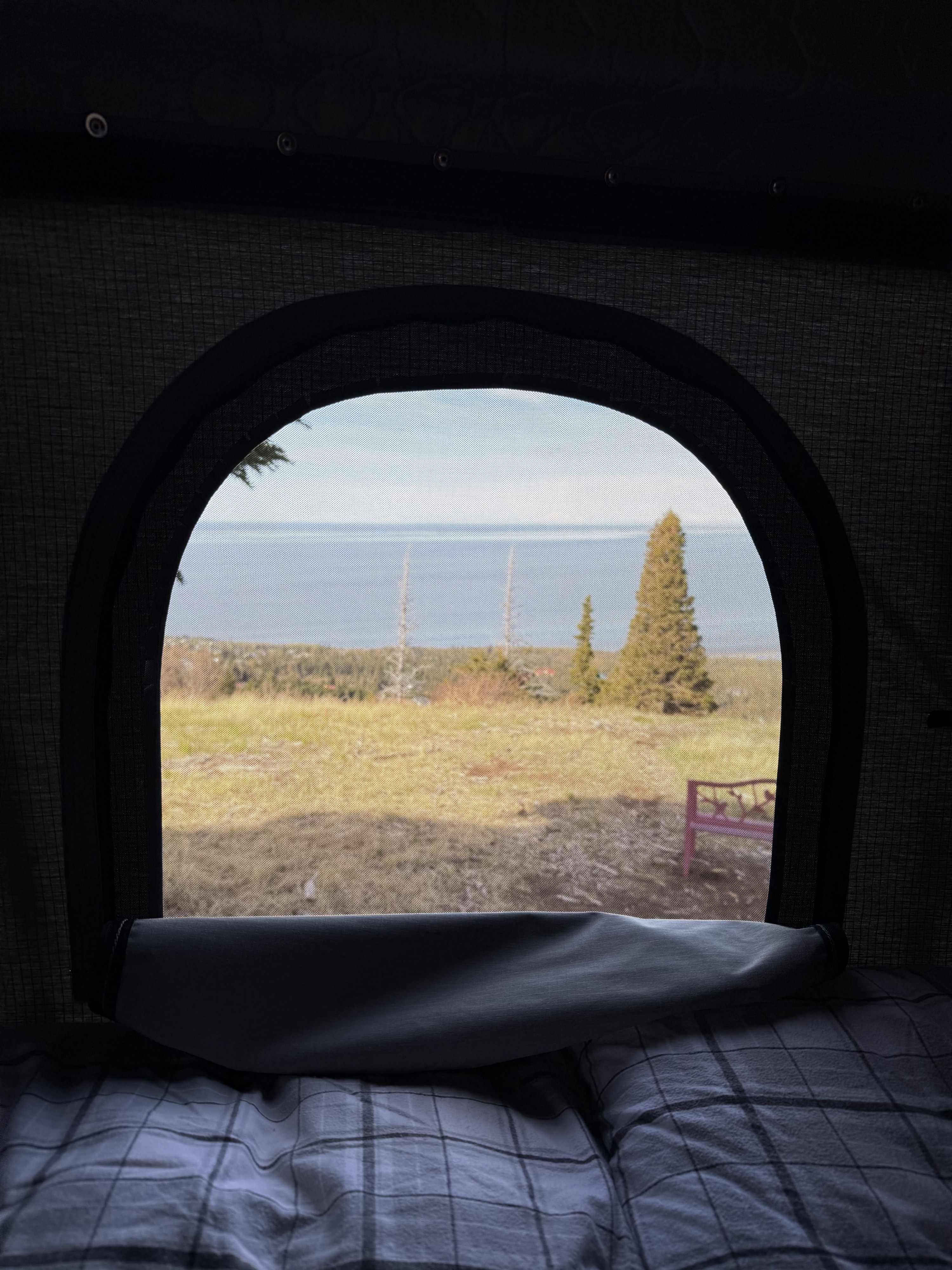 Comfortable RV rental: plaid bedding & a screened window reveal a scenic outdoor view of a field, trees, and water.. Dodge Grand Caravan 2017