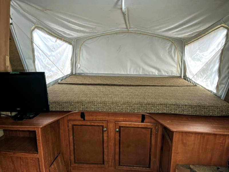 Inside a comfortable RV rental, featuring a wide bed with a patterned mattress. A TV sits on adjacent wooden cabinets, perfect for travel.. Jayco Jay Feather Ultra Lite 2013
