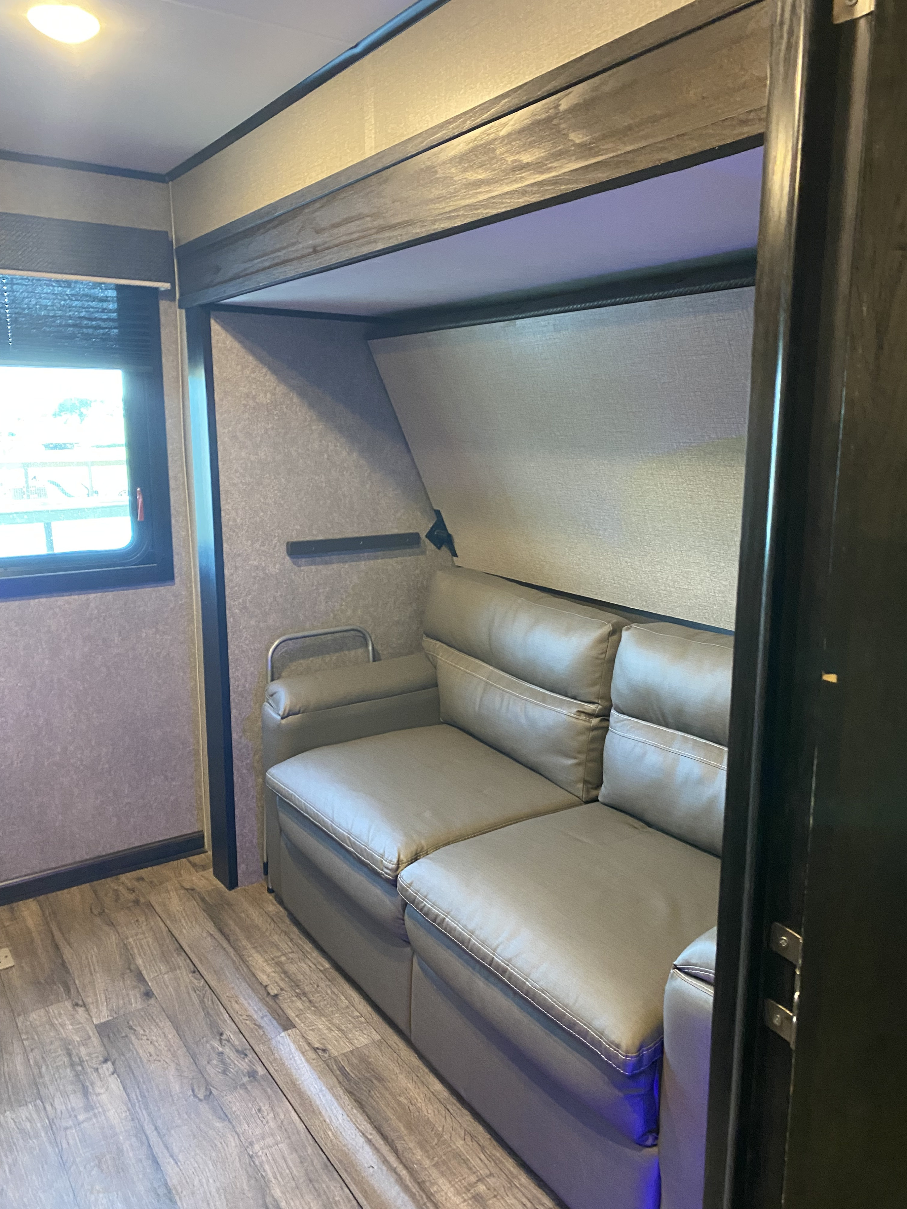 This RV rental features a cozy living space with a grey sofa, wood-look flooring, and a window with blinds. Ideal for travel.. Jayco Eagle HT 2021