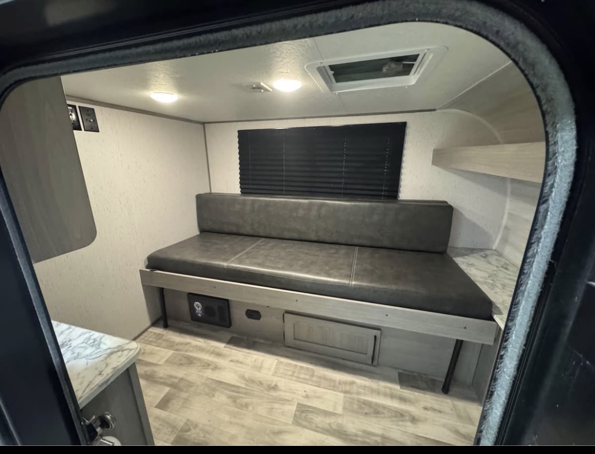 View into an RV rental's compact interior: a bench/bed, window with blinds, overhead lights, storage, and small counter.. Coleman Rubicon 1200RK 2023