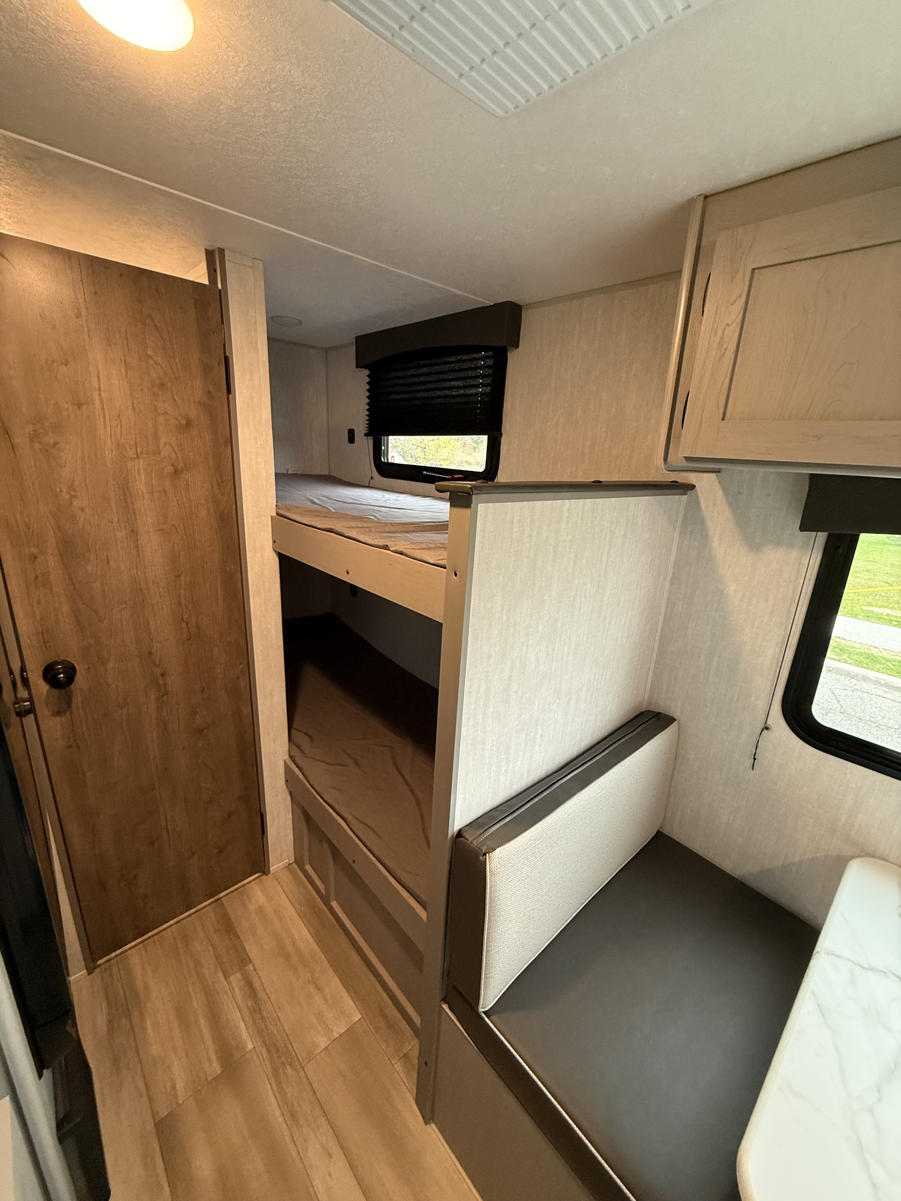 RV rental interior: light wood-look floor, bunk beds with gray bedding, a gray and light dinette, and a window with blind.. Coachmen Clipper 17CBH 2025