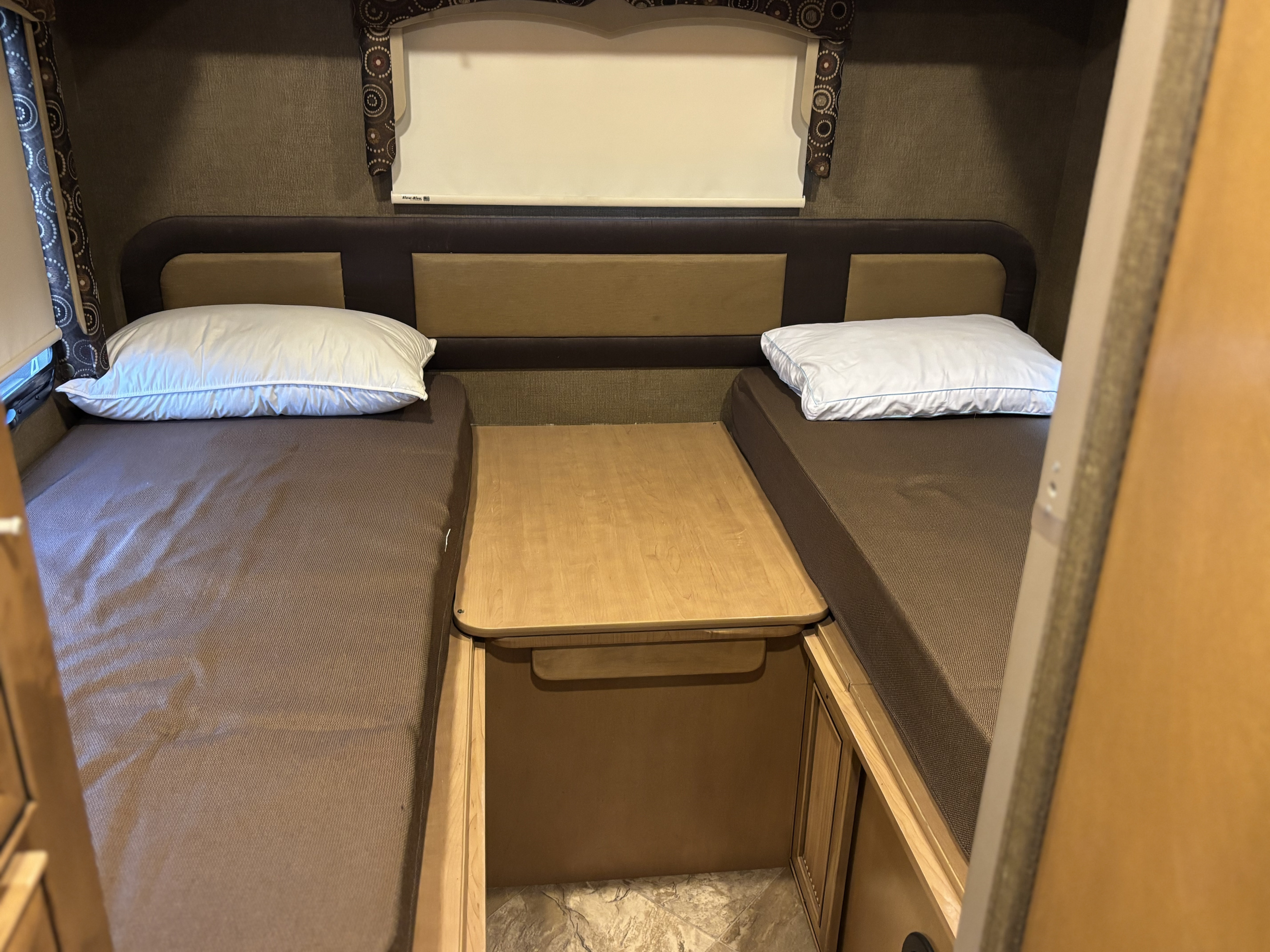 RV rental interior showing two brown-covered twin beds with white pillows, separated by a wooden table, under patterned curtains.. Thor Siesta 2016
