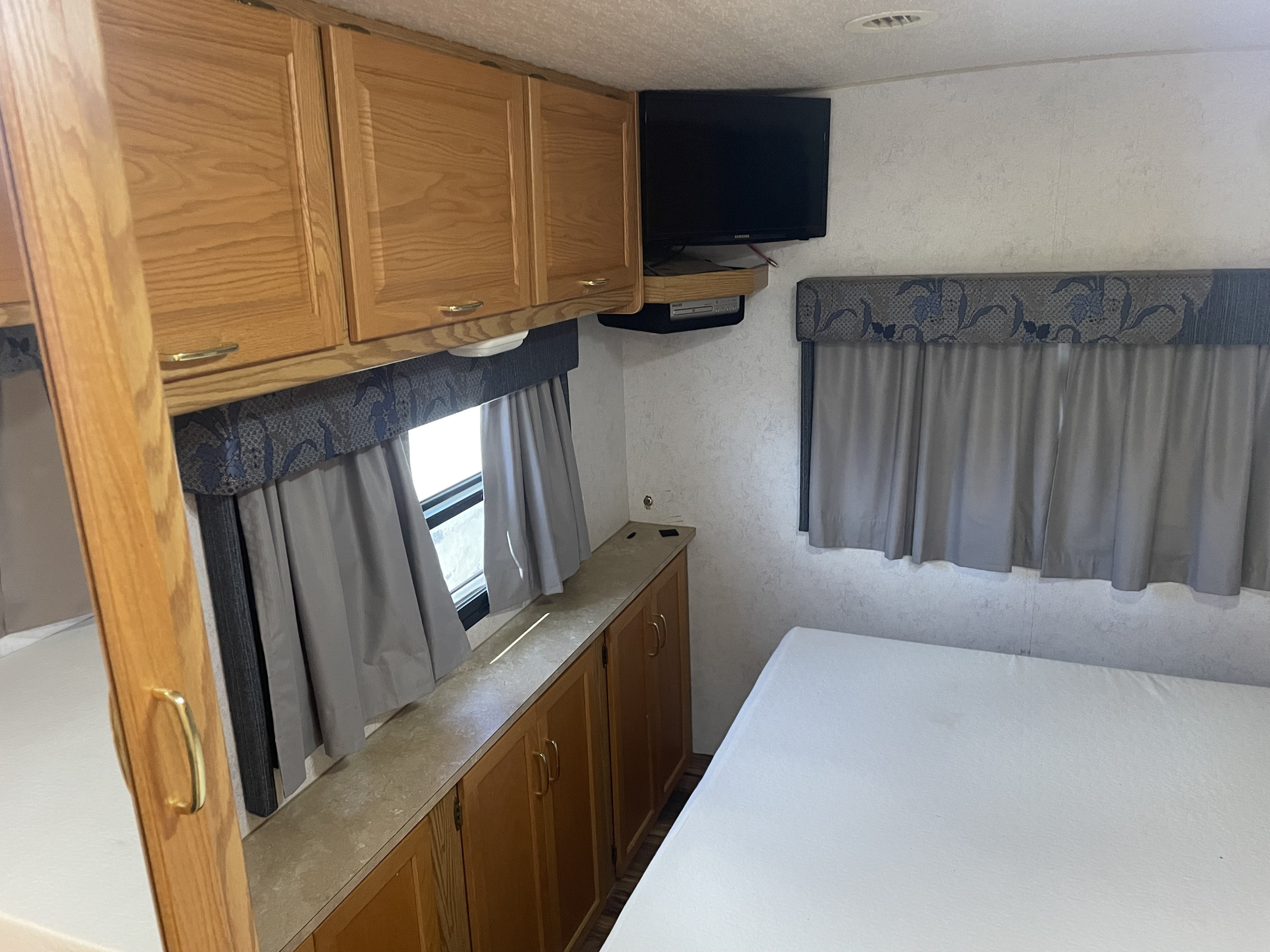 RV rental interior: light wood cabinets, wall-mounted TV, comfy bed, and windows with grey curtains.. Itasca Spirit 2003