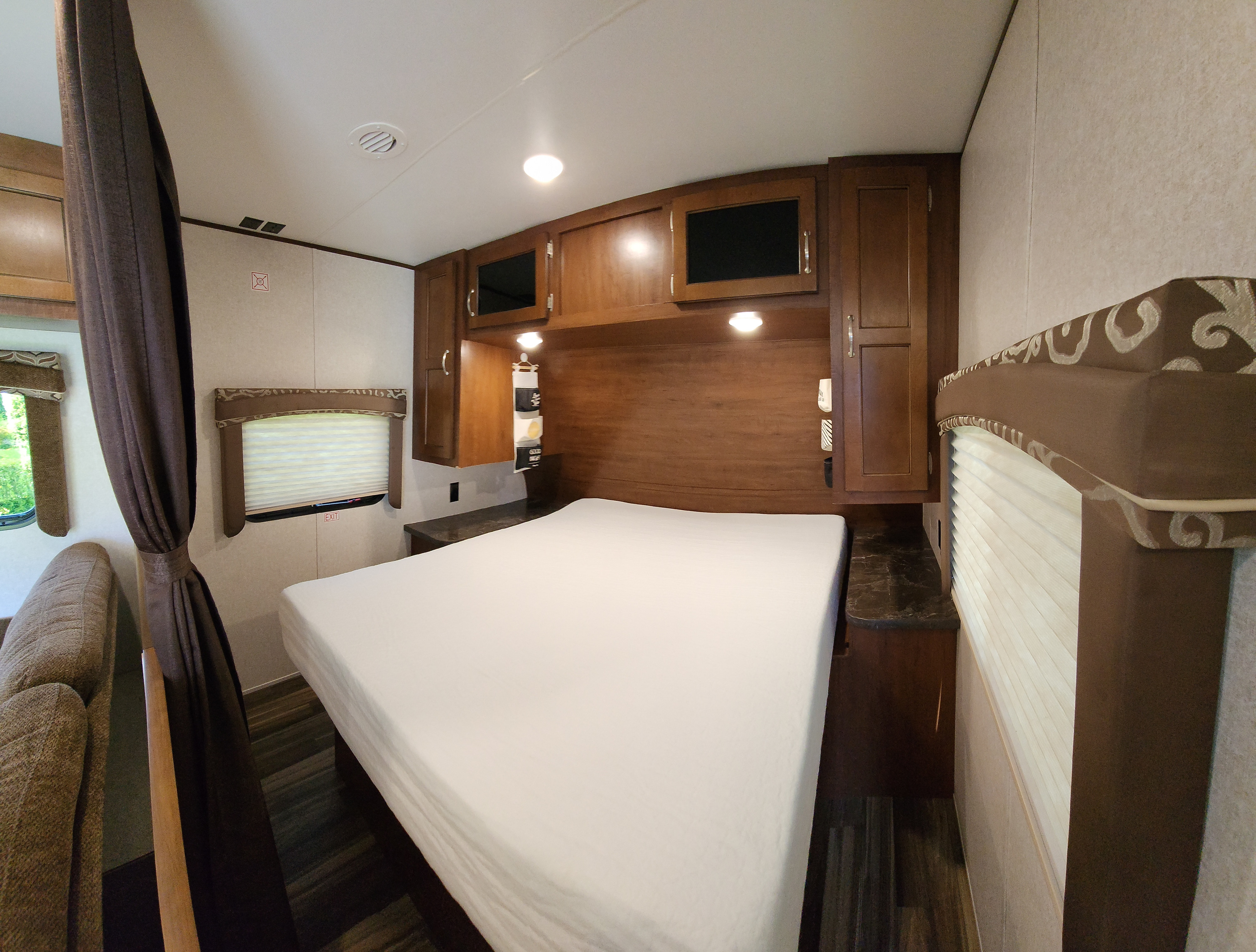 RV rental bedroom view, showing a large white-sheeted bed, wood overhead cabinets, and two bright windows.. Jayco Jay Flight 2017