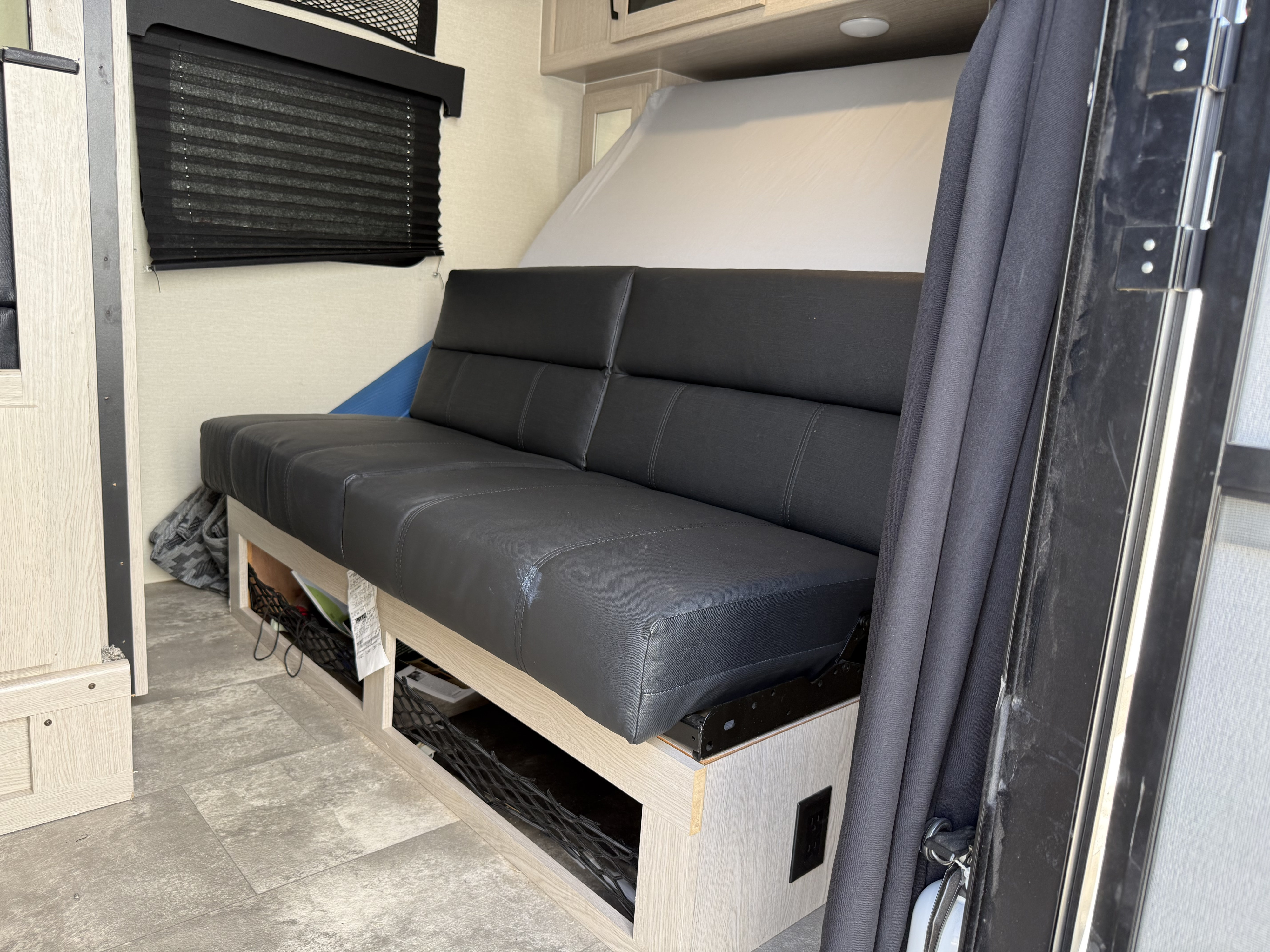 RV rental interior: black futon with under-seat storage, pleated window blind, and fold-up bed. Compact living space.. Coachmen Apex Nano 2022