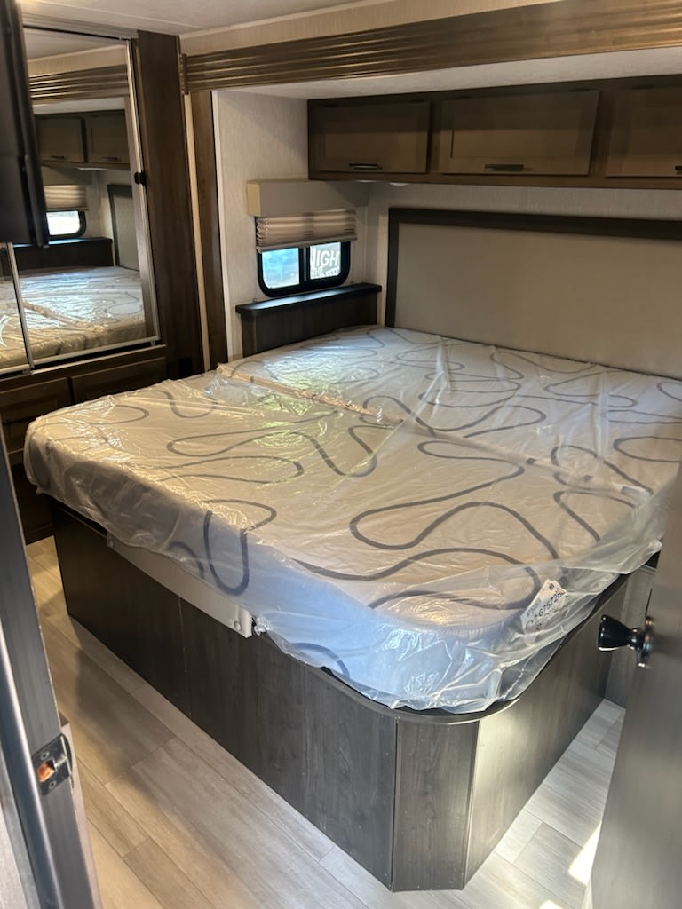 Inside an RV rental bedroom: a new, plastic-wrapped mattress on a queen bed base, with storage cabinets above and a window.. Forest River Coachmen Leprechaun 2023