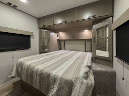 Cozy RV rental bedroom with a comfortable striped bed, built-in overhead cabinets, and soft lighting. Perfect for your next adventure.. Grand Design Reflection 2024