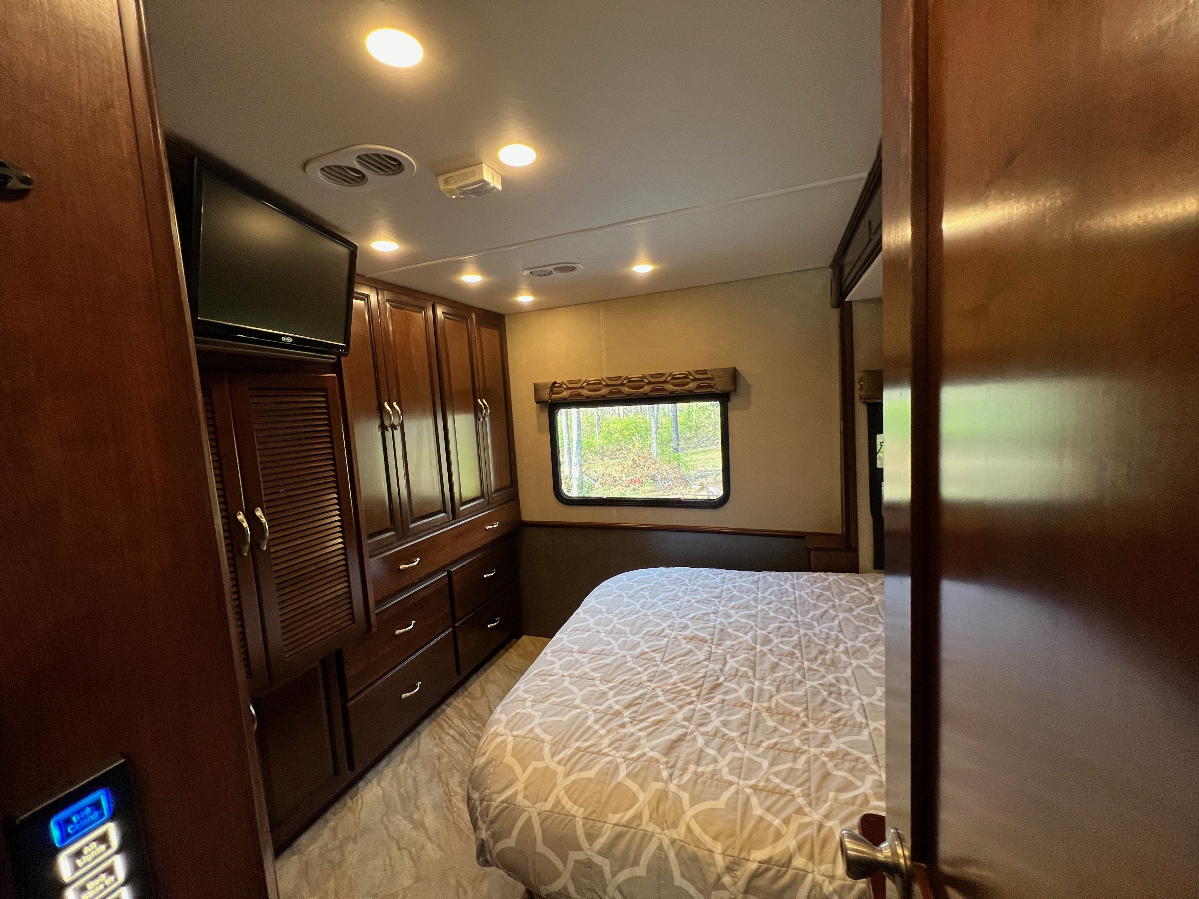 Cozy RV rental bedroom displaying a patterned bed, dark wood cabinets, TV, and a window with a treed view.. Fleetwood Bounder 2017