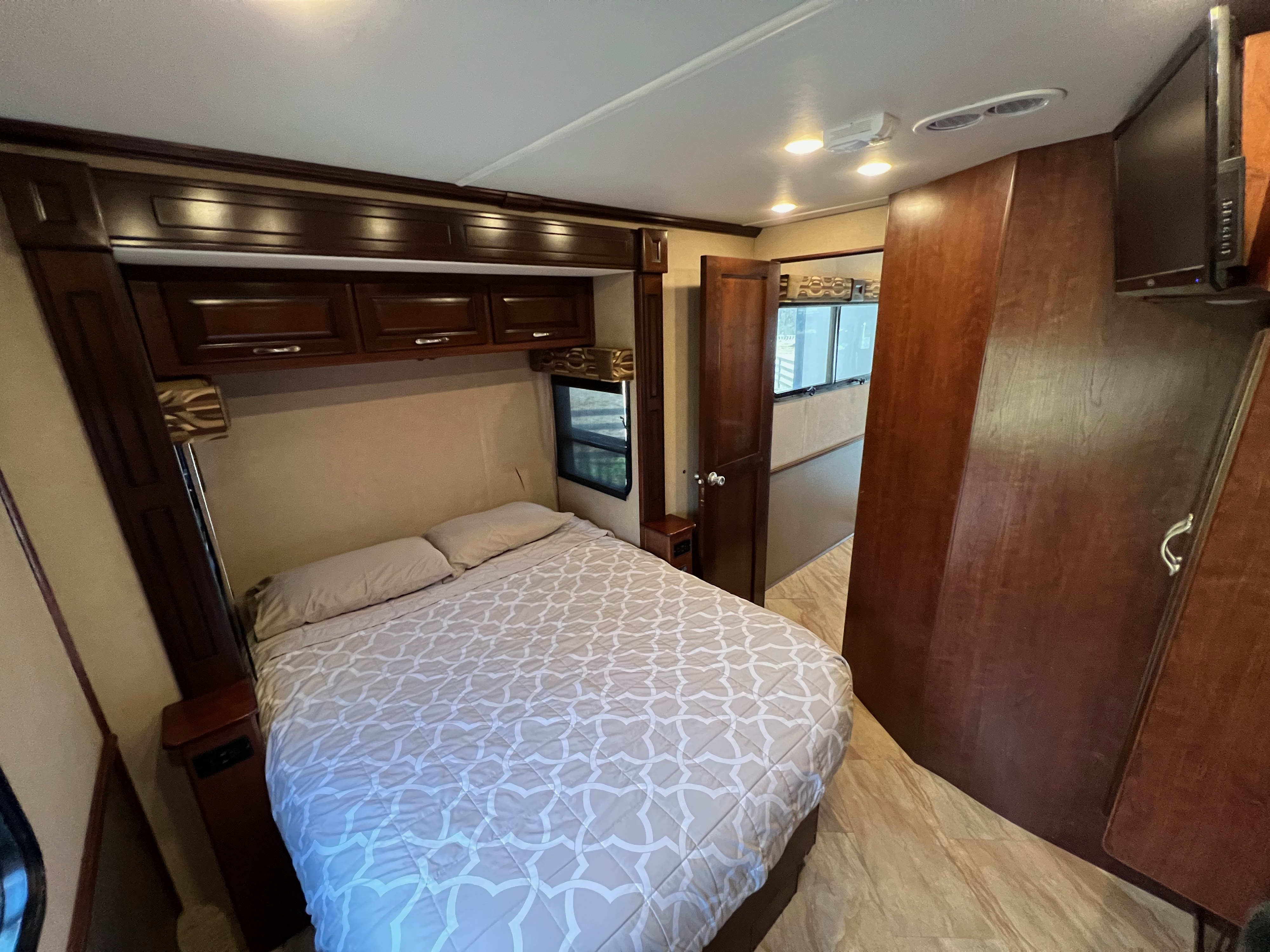Inside an RV rental: a compact bedroom with a patterned bedspread, overhead cabinets, and a mounted TV.. Fleetwood Bounder 2017
