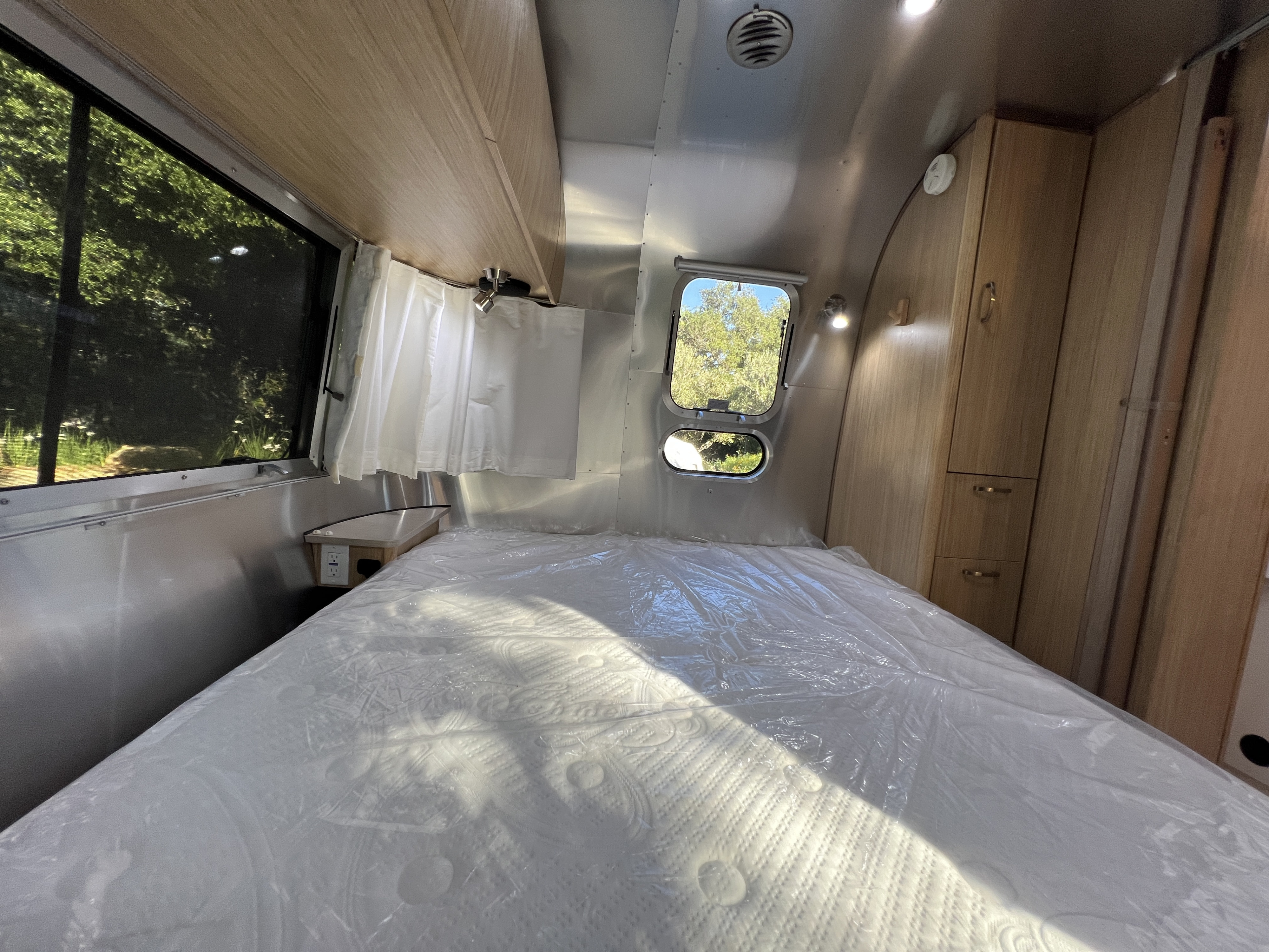 RV rental bedroom: new, plastic-wrapped mattress, curved aluminum walls, wooden cabinets, and bright windows.. Airstream Flying Cloud