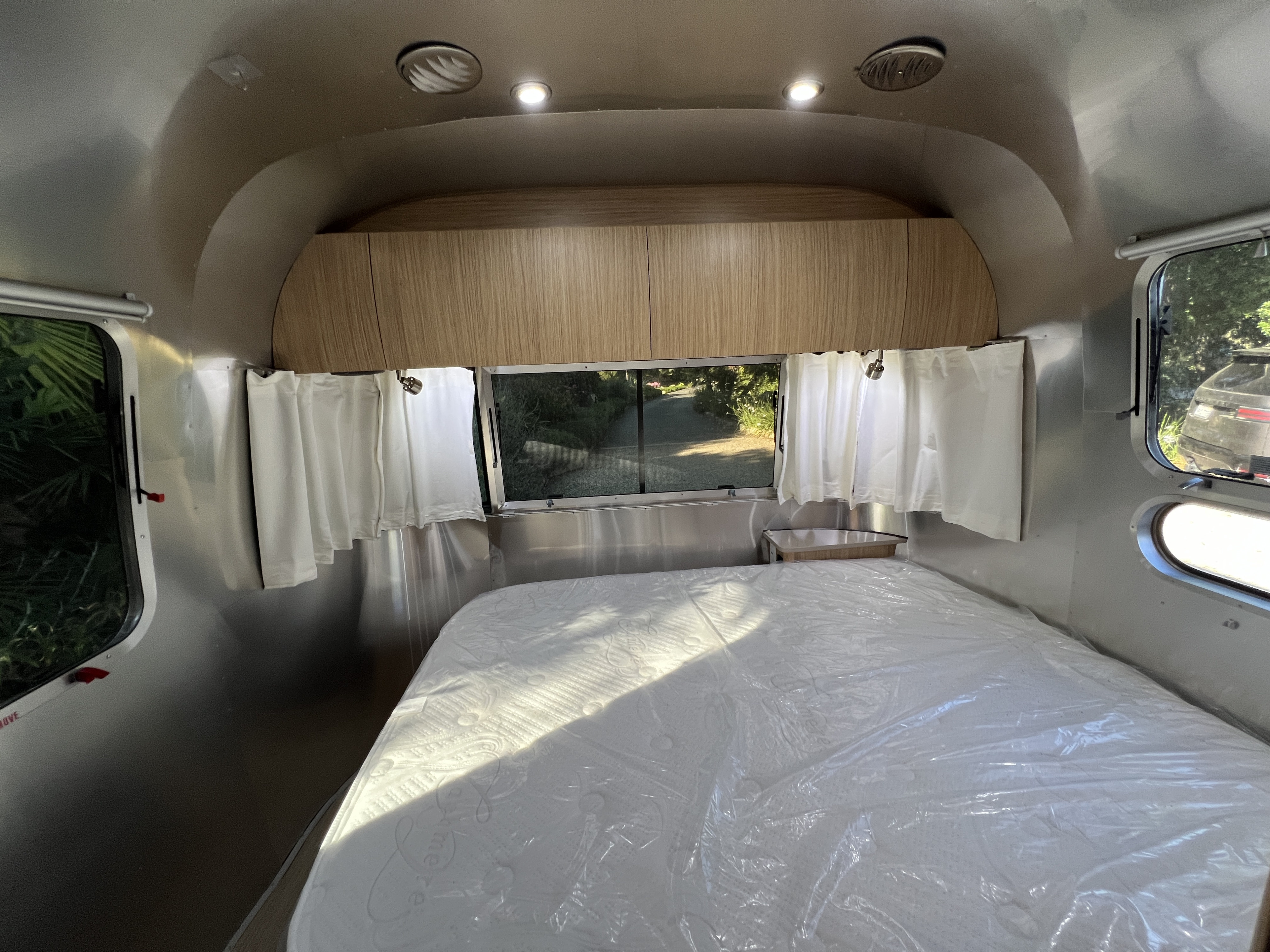 Airstream RV rental bedroom with a clean, plastic-wrapped mattress, wood cabinets, and windows with white curtains.. Airstream Flying Cloud