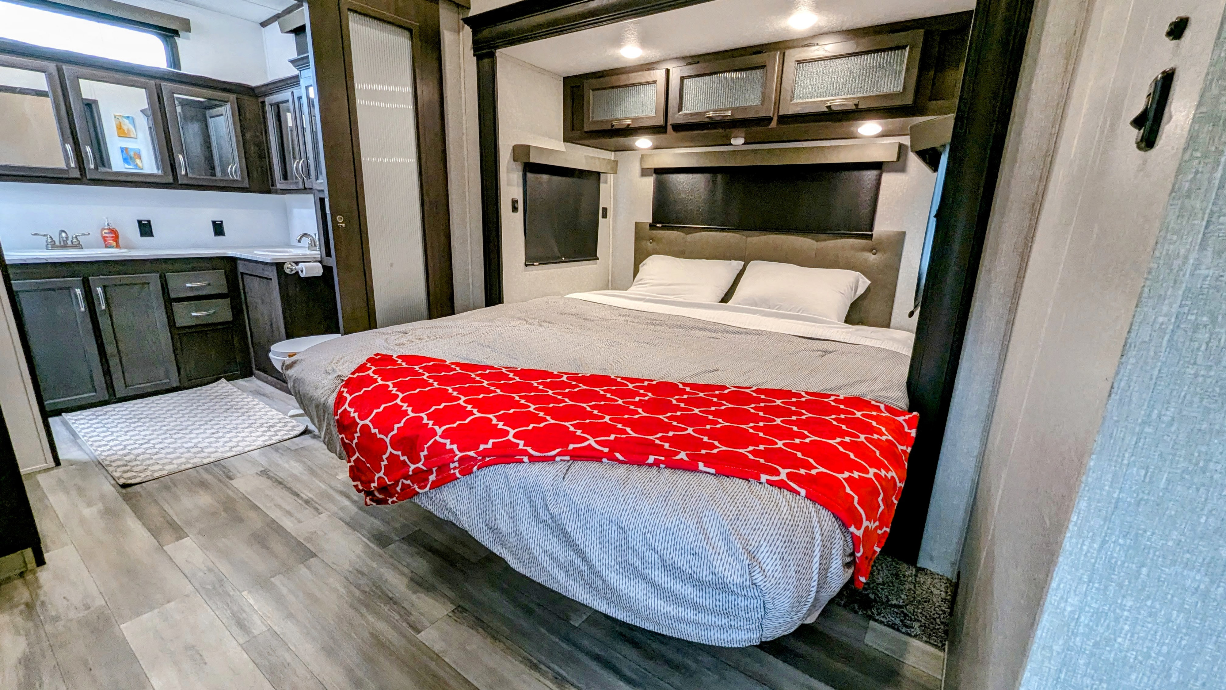 Comfortable RV rental bedroom with queen bed & red blanket. Modern overhead cabinets and a glimpse of the compact kitchen.. Forest River Brookstone 2022
