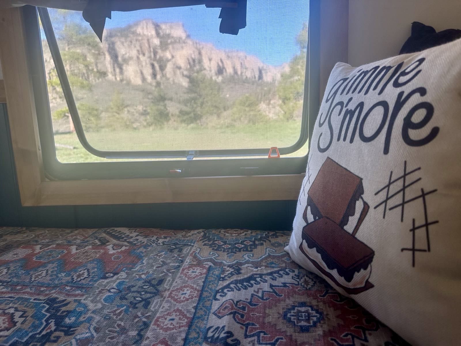Inside a cozy RV rental: a window with mountain views, a s'mores pillow, and patterned bedding.. Heartland RVs Prowler 2022