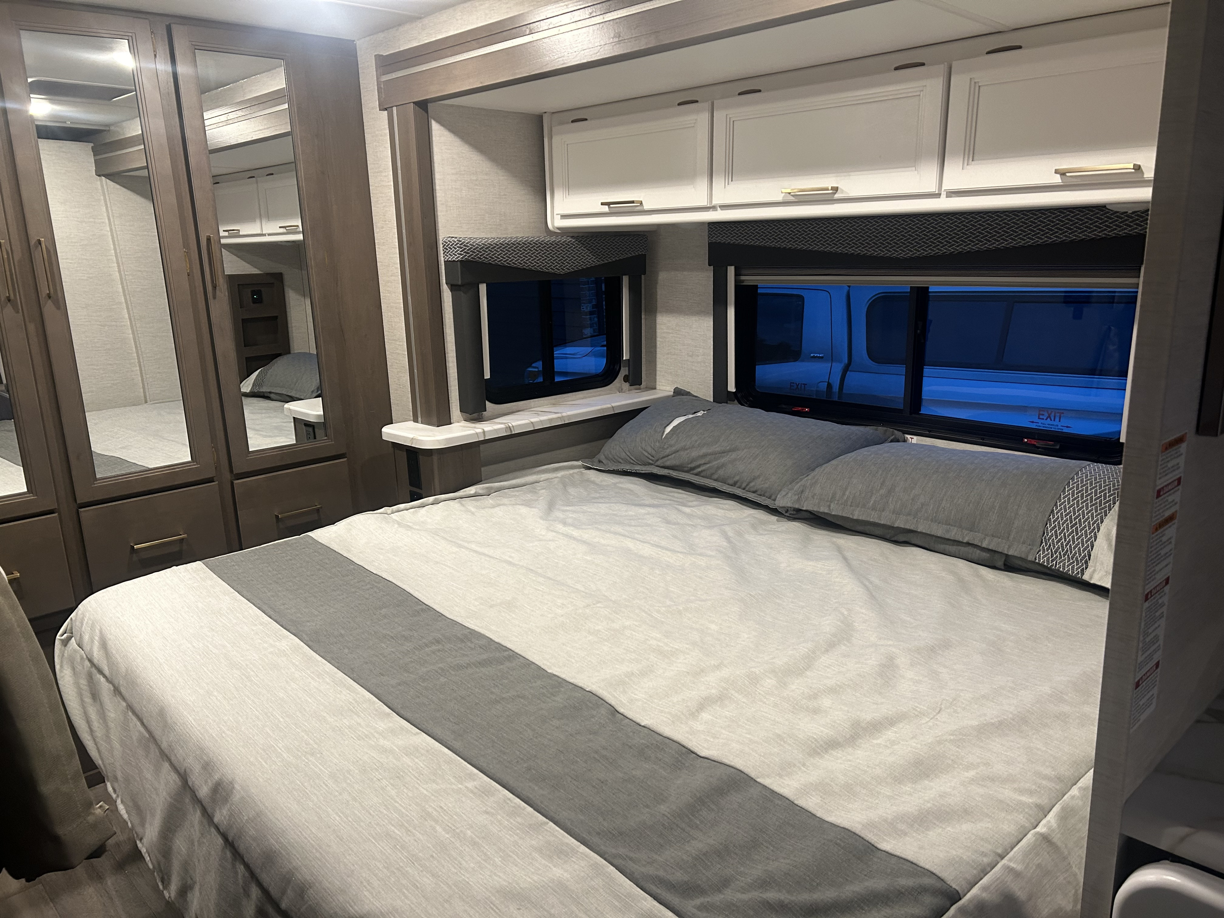 Modern RV rental bedroom featuring a light grey bed, mirrored wardrobe, and white overhead cabinets.. Thor Four Winds 2024