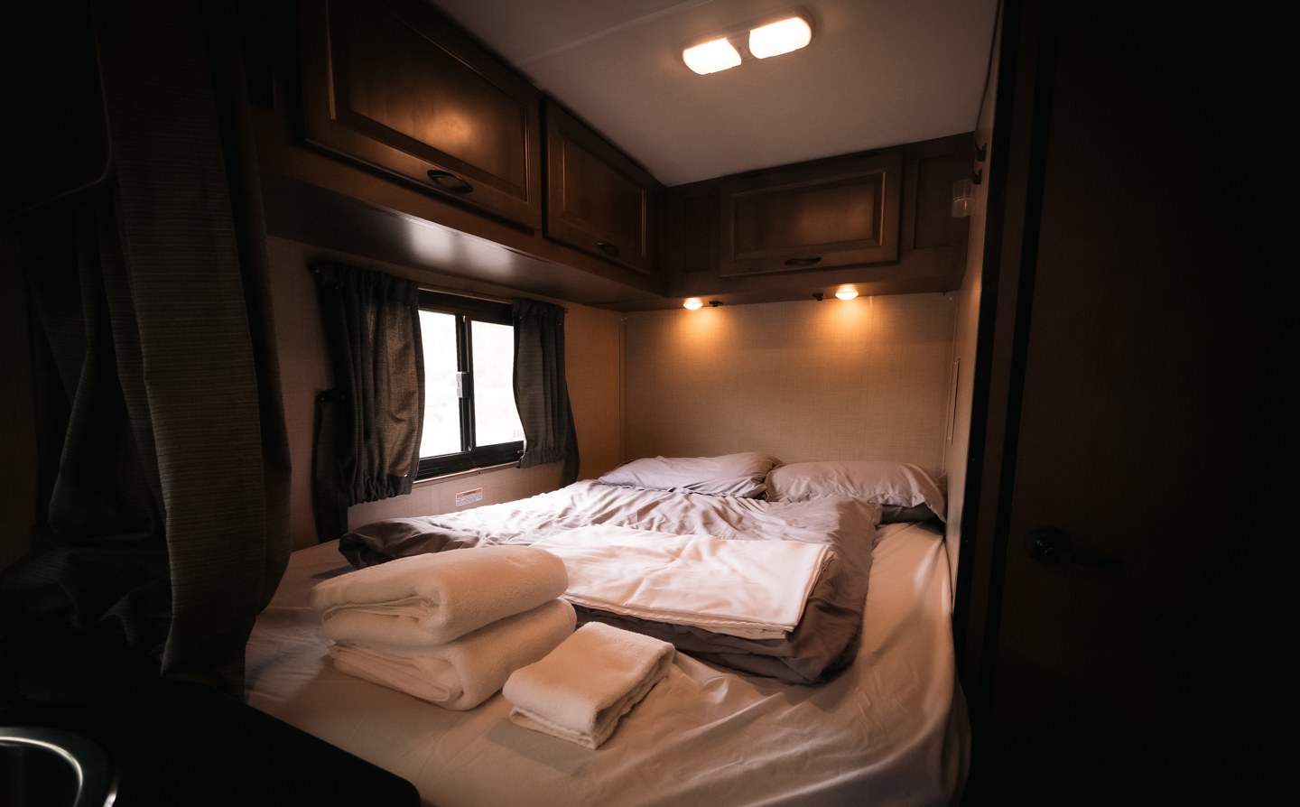 Inside an RV rental, this cozy bedroom features a bed with grey and white linens, overhead storage, and a window.. Wayfarer Wayfarer Motorhome 2020