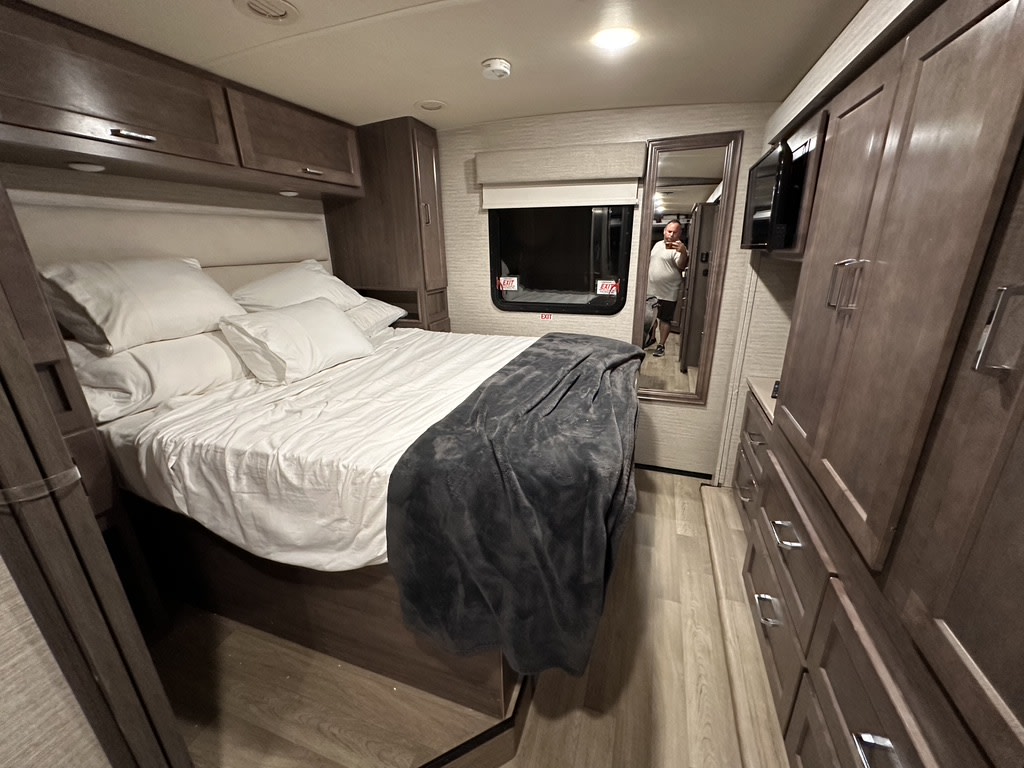 Cozy RV rental bedroom with queen bed, white bedding, built-in storage, mirror, and TV. Ready for travel.. Winnebago Adventurer 2021