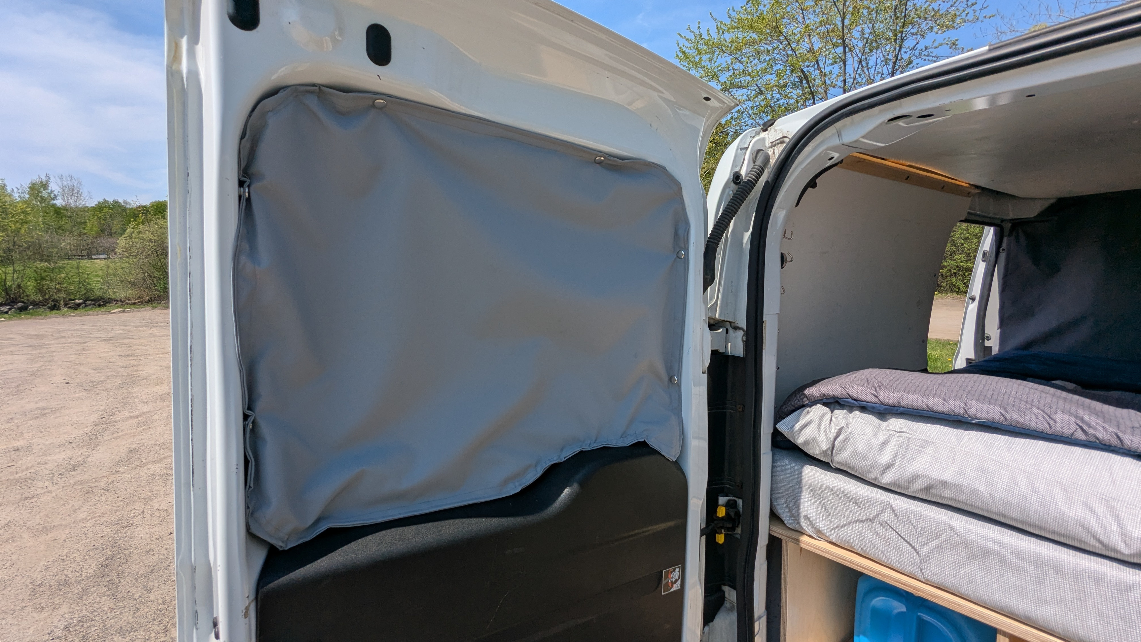 Open rear doors of an RV rental van reveal a comfortable bed, wood framing, and custom gray paneling for the camper interior.. Dodge Promaster City 2015