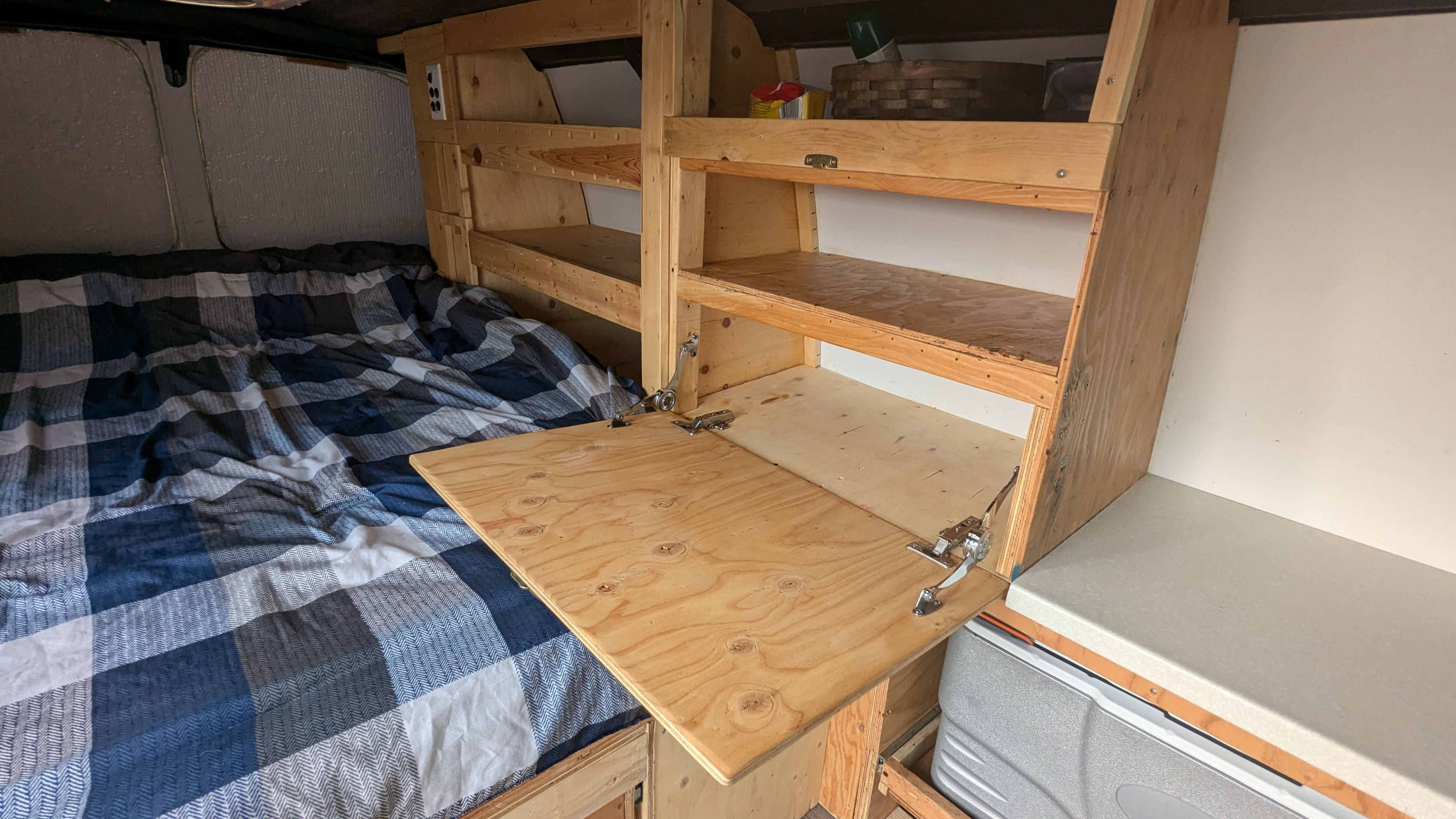Inside an RV rental: plaid bed, custom wood shelving, and a hinged, foldable table. Efficient camper van living space.. Gmc Savanna 2015