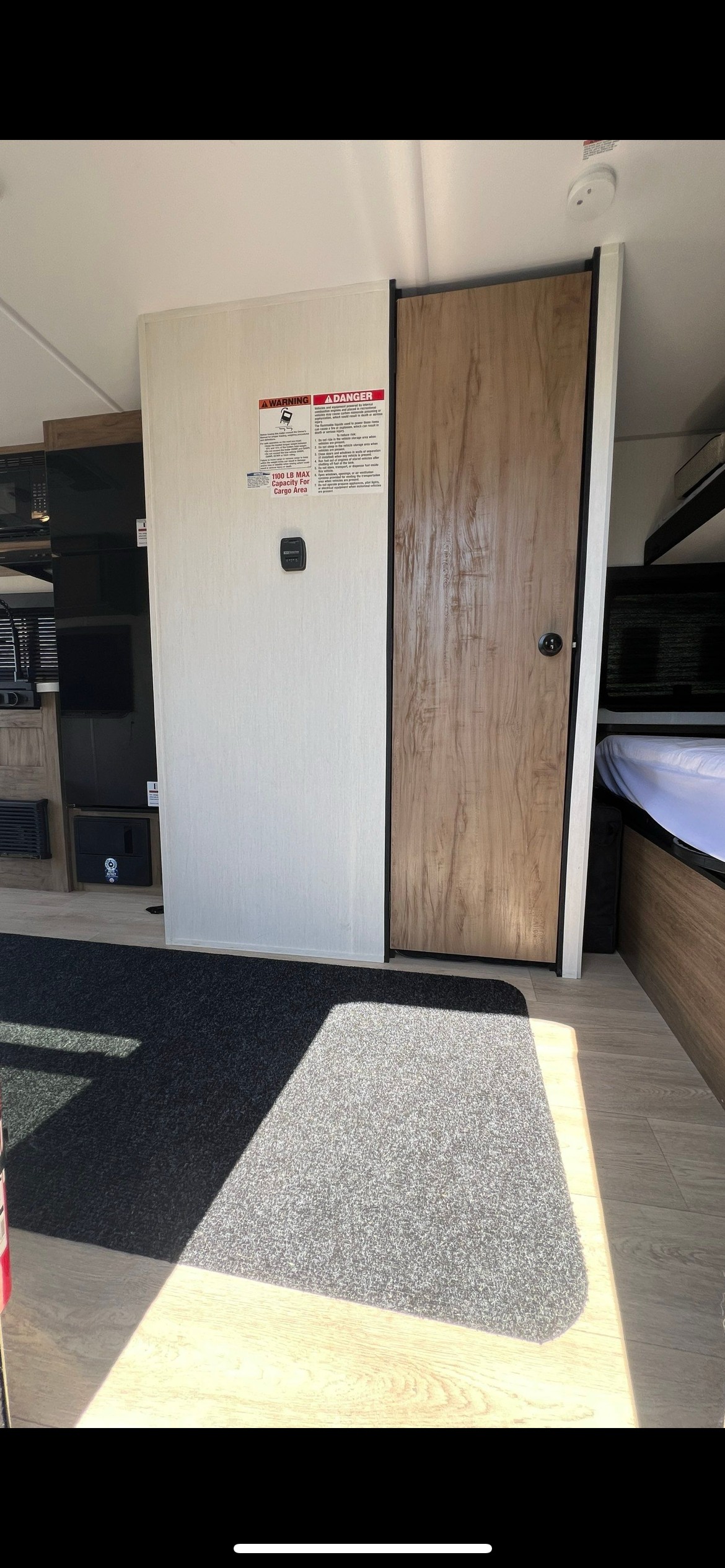 RV rental interior: View of cargo area wall with warning labels, wooden door, and part of a bed. Carpeted floor with sunlight.. Forest River Wolf Pup 2025