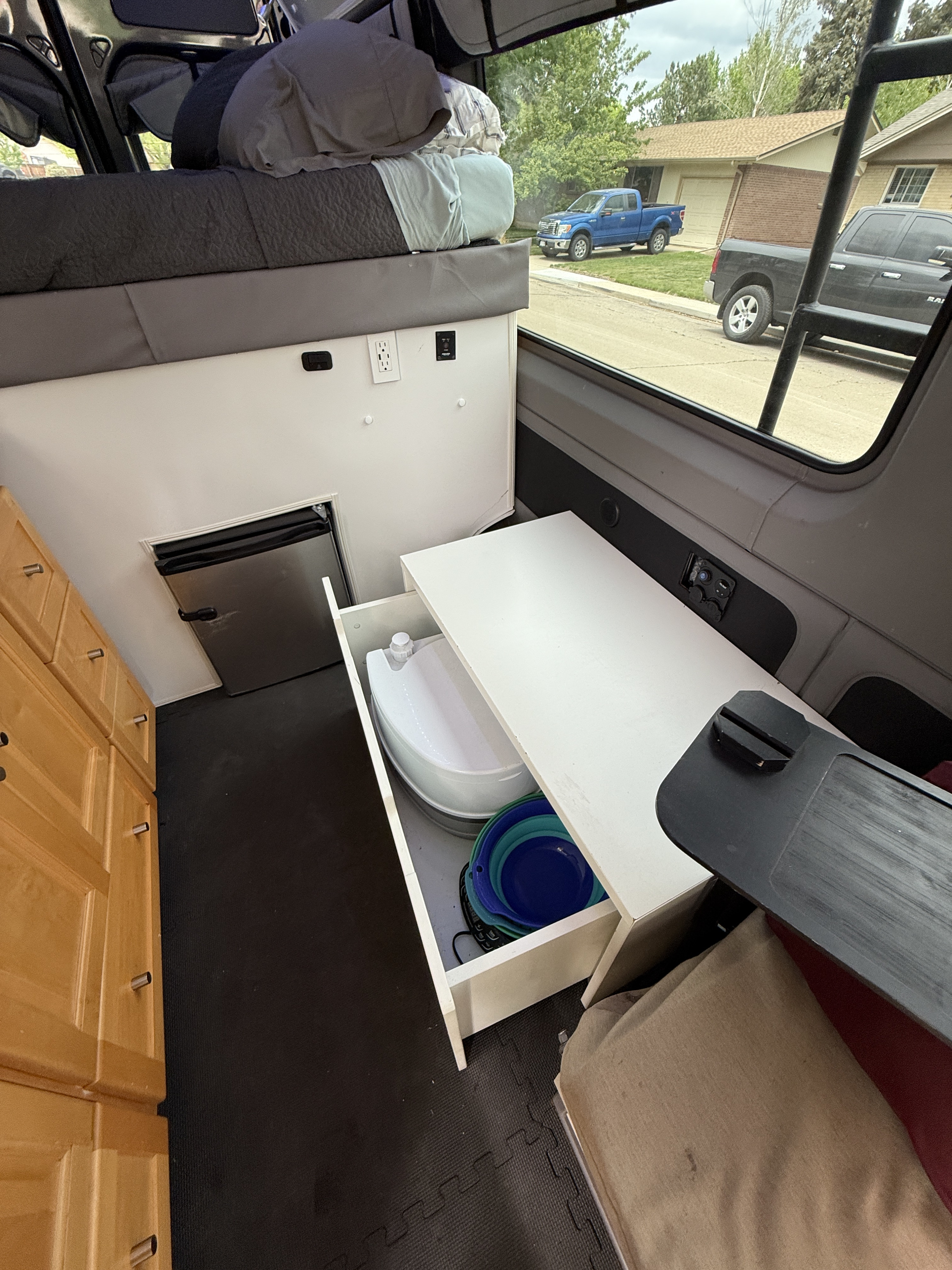 This RV rental camper van showcases a compact interior with an elevated bed, small fridge, pull-out toilet, and cabinets.. Mercedes-Benz Sprinter 419 LWB High Roof 2015