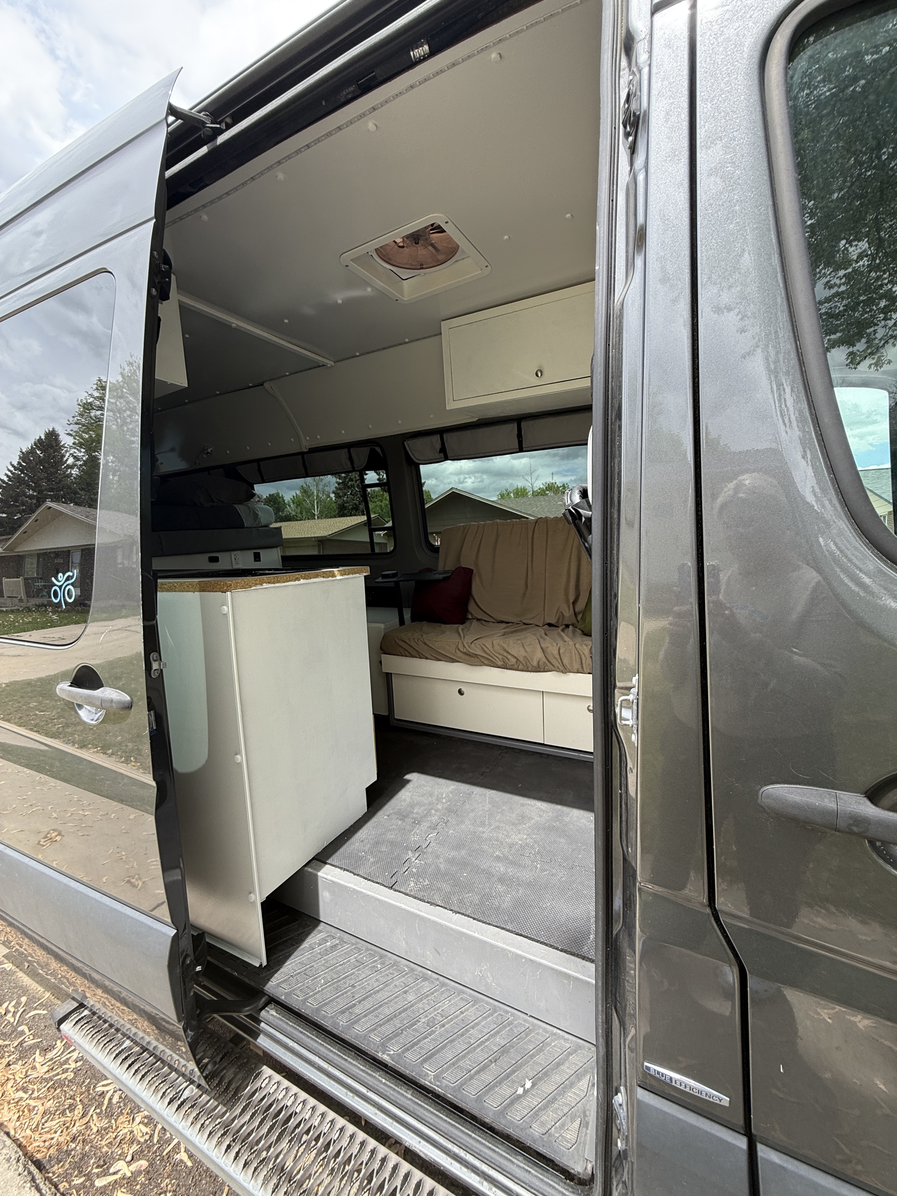 RV rental: Interior view of a grey converted camper van through its open side door, revealing a bed, storage, and ceiling fan.. Mercedes-Benz Sprinter 419 LWB High Roof 2015