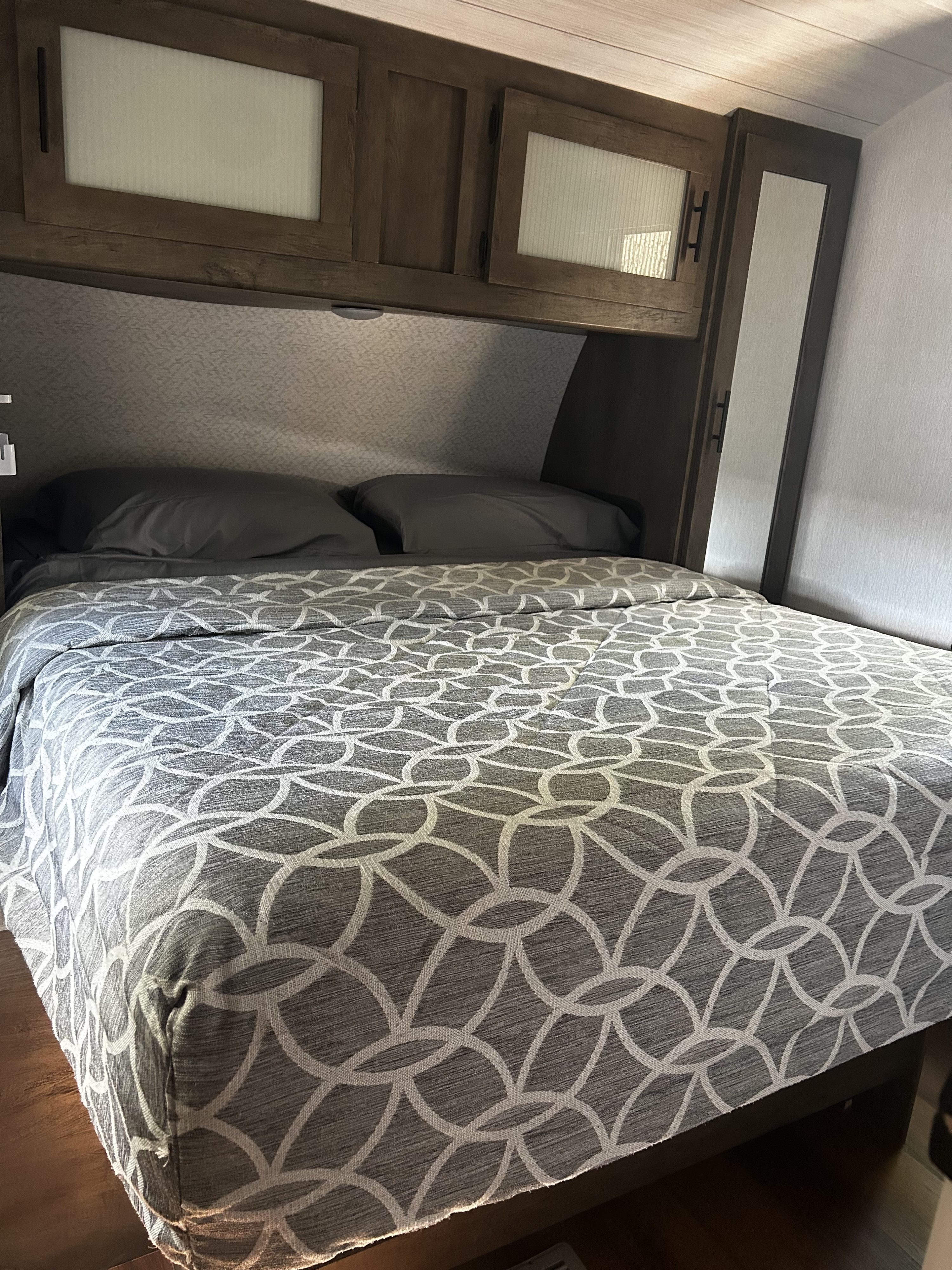 This RV rental bedroom features a comfy bed with gray patterned comforter & pillows, plus overhead wooden cabinets.. Forest River Wildwood 2022