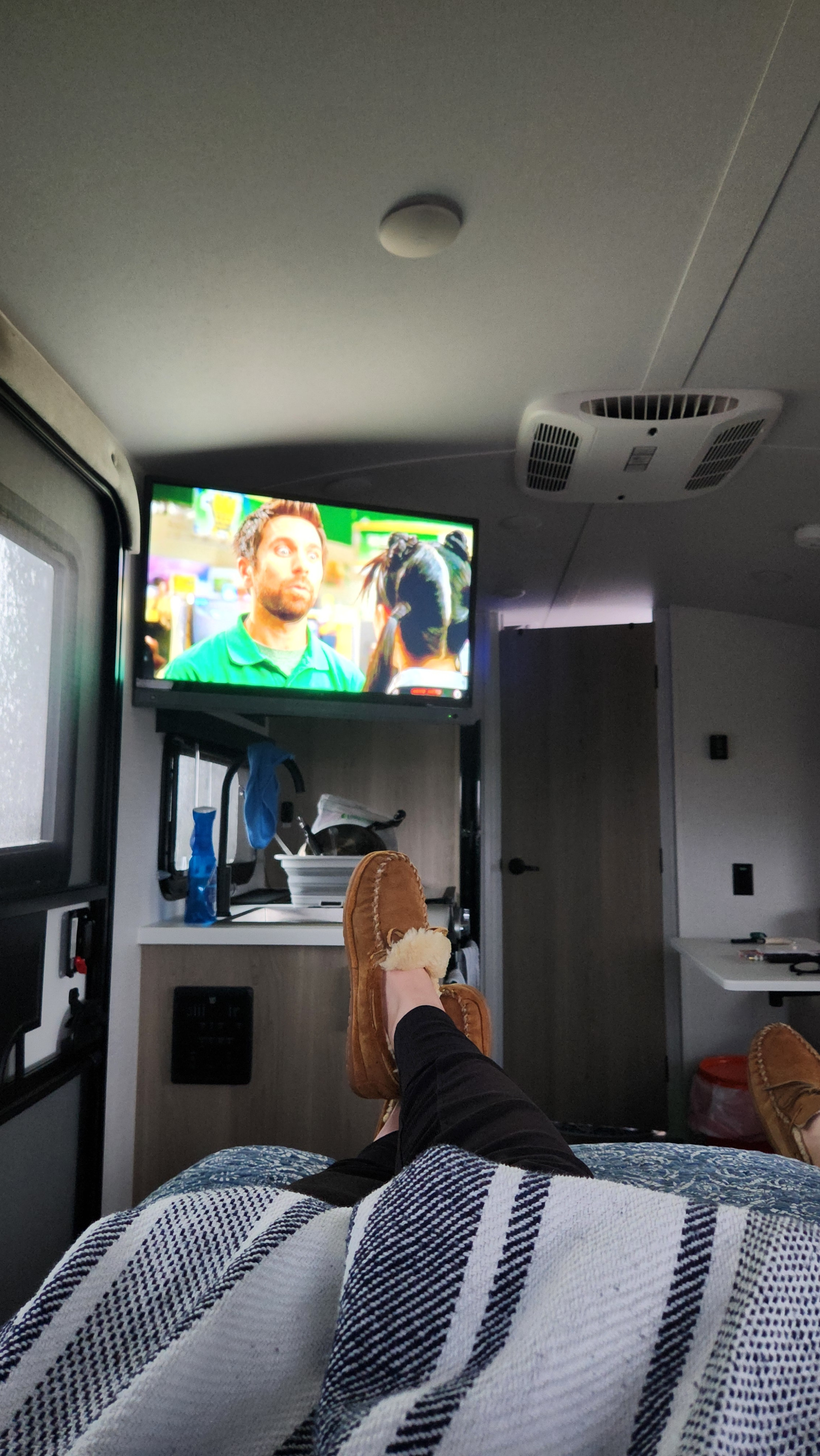 Inside an rv rental, a person relaxes with feet in brown moccasin slippers on a striped blanket, watching TV.. Forest River Rockwood Geo Pro 2024