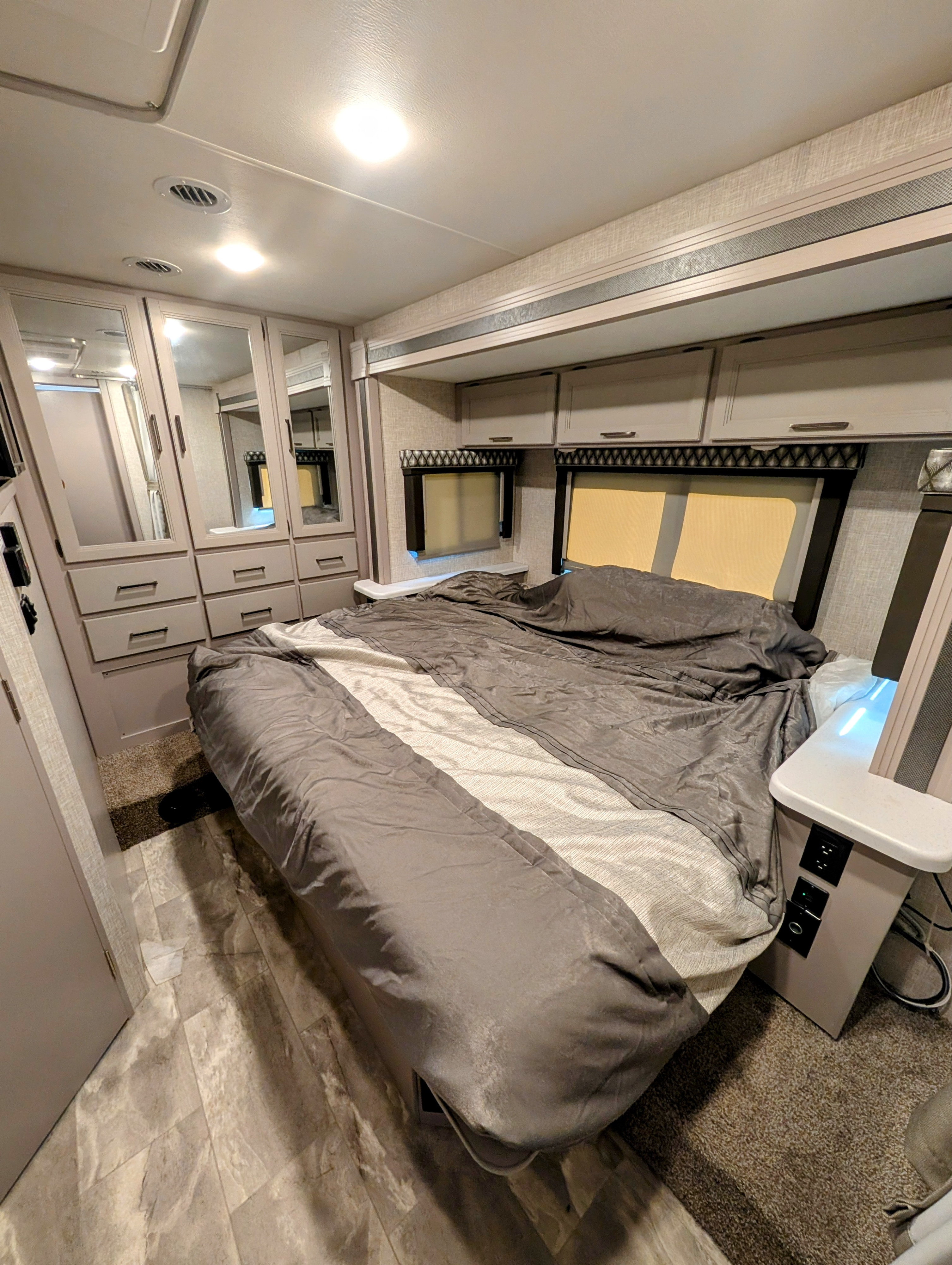 Relax in this comfortable RV rental bedroom, featuring a queen bed, mirrored wardrobes, windows, and overhead storage.. Thor Quantum 2022
