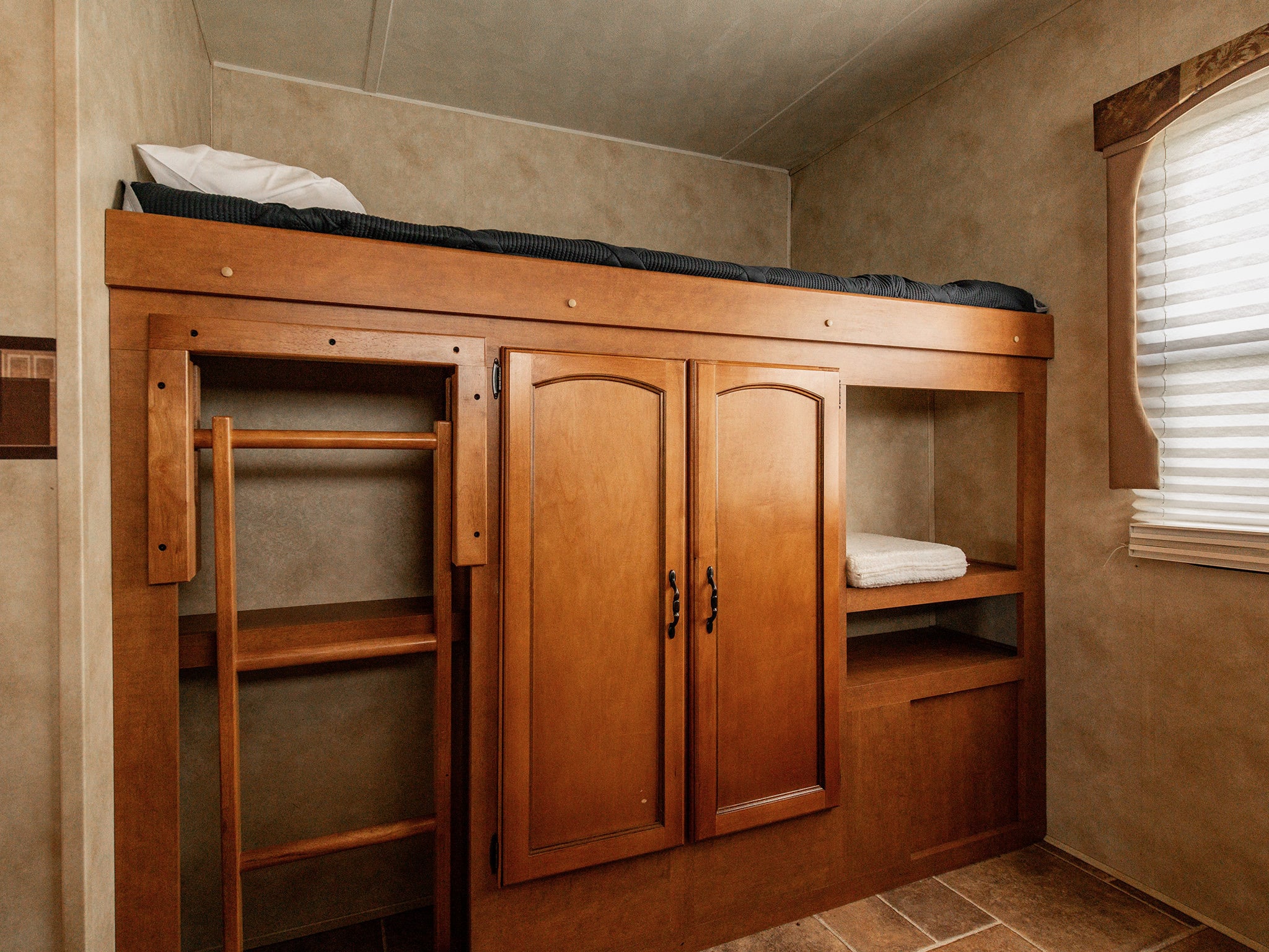 RV rental bunk bed with mattress, ladder, cabinets, and open shelving for storage. Bright window with blinds.. Forest River Palomino Puma 2012