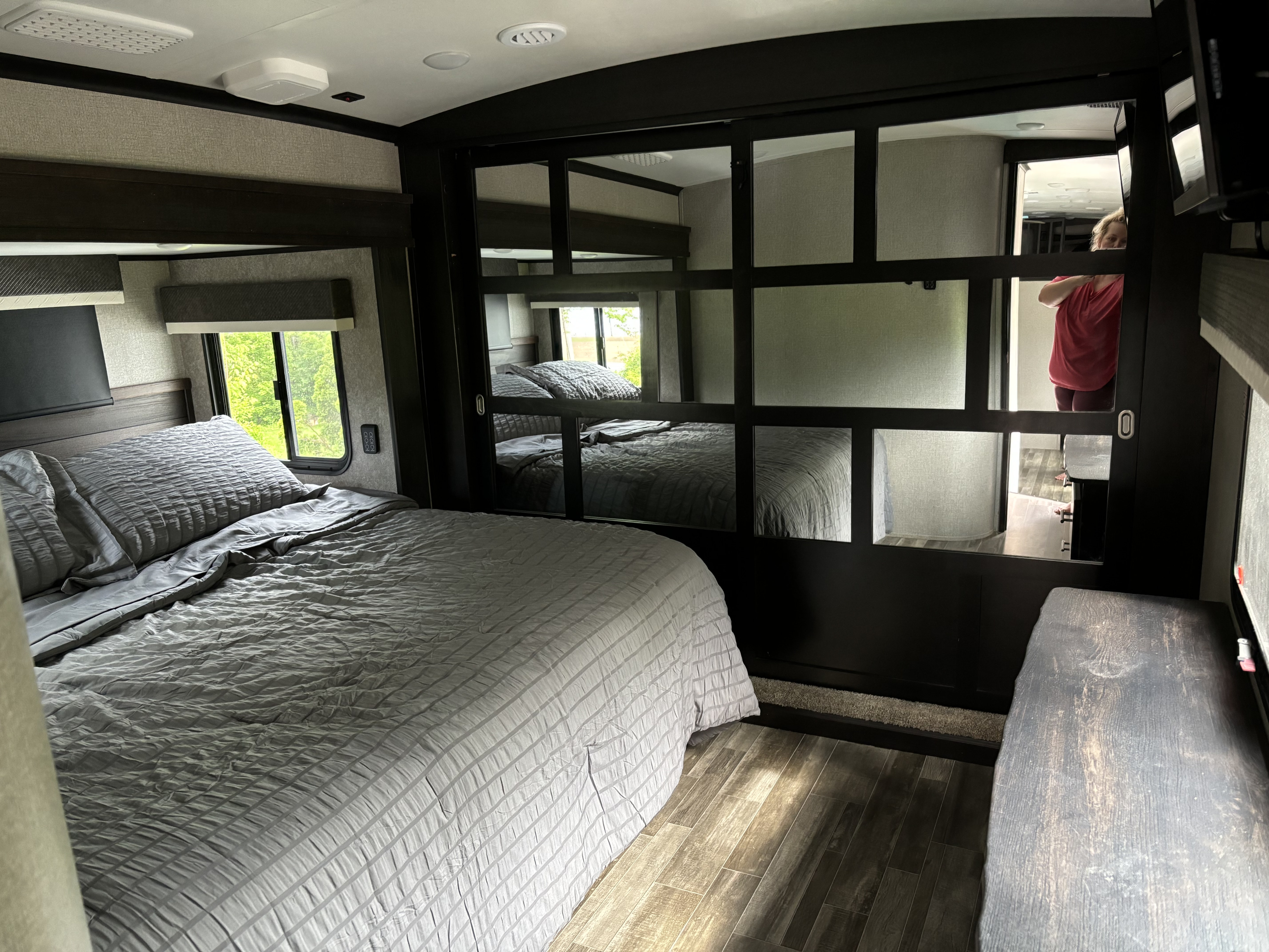 Modern RV rental bedroom with comfy queen bed, grey bedding, mirrored wardrobe door reflecting a person. Wood floor.. Jayco North Point 2021