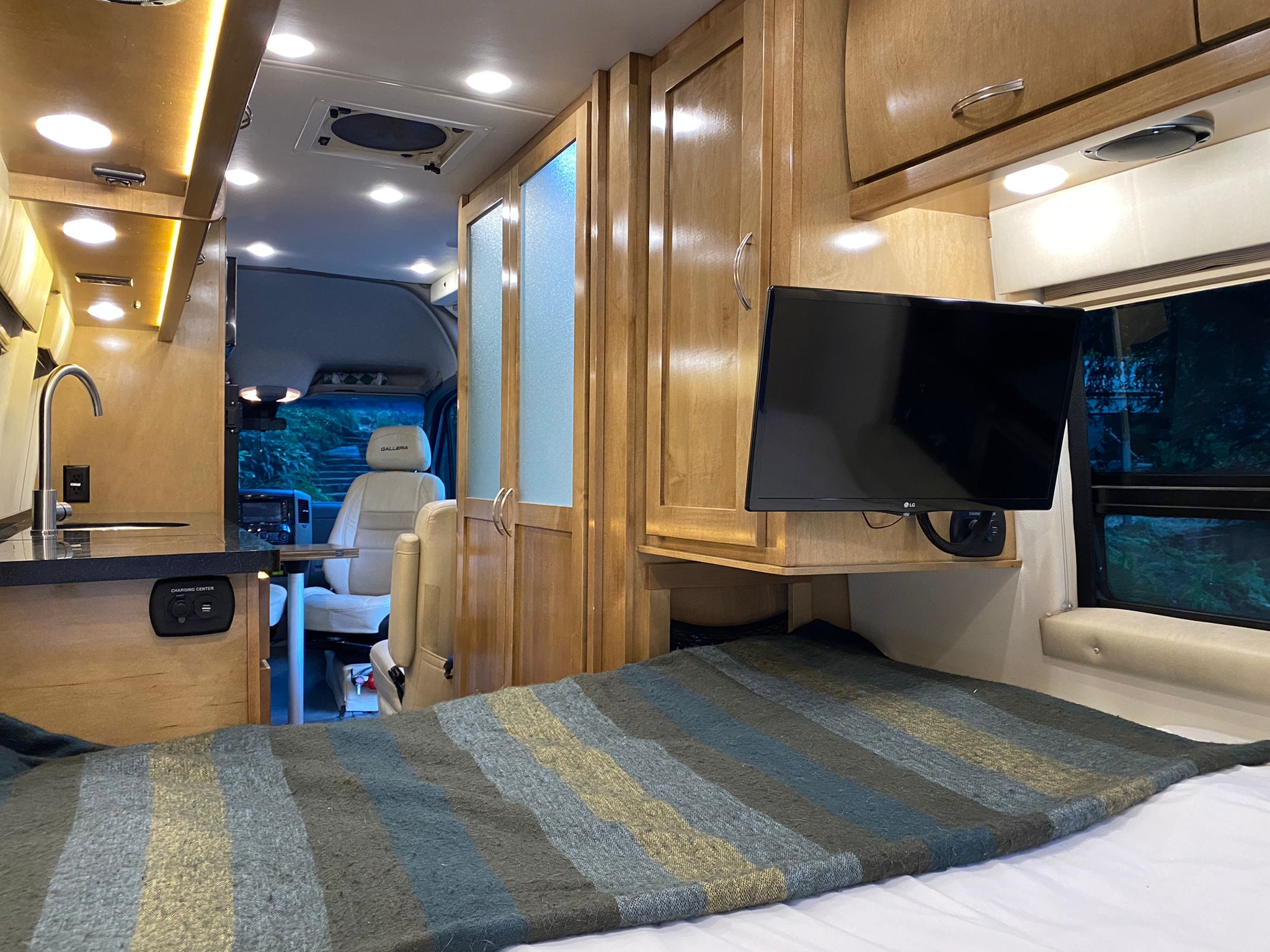 Bright and modern RV rental interior showing a bed with a striped blanket, kitchen, wood cabinets, and mounted TV for a comfortable journey.. Mercedes Benz Sprinter/ V6 3322 19 GALLERIA 24T RV Brilliant Silver 2018