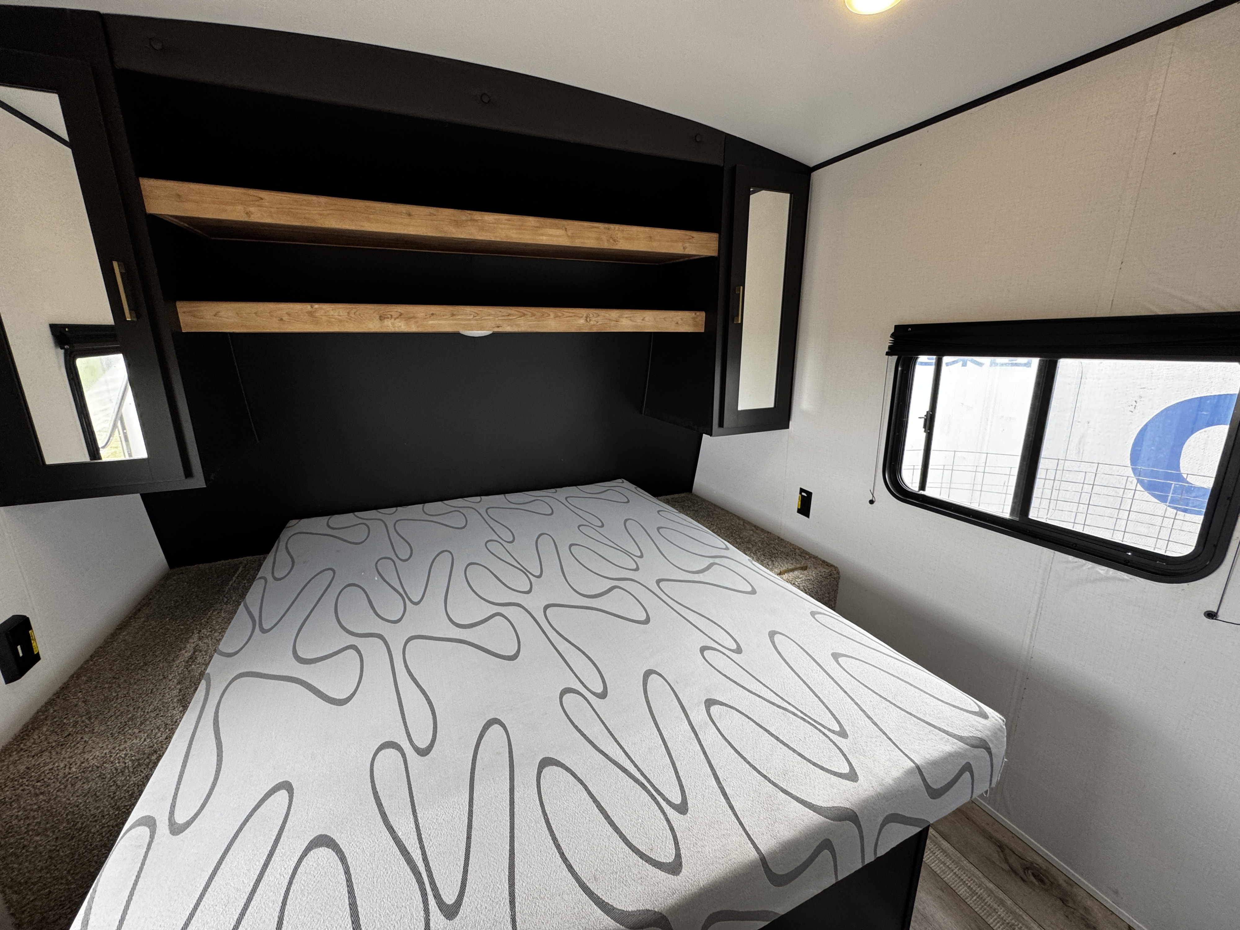 Get comfy in this RV rental bedroom: bed with patterned cover, black overhead storage, side mirrors, and window.. Keystone RV Springdale 2023