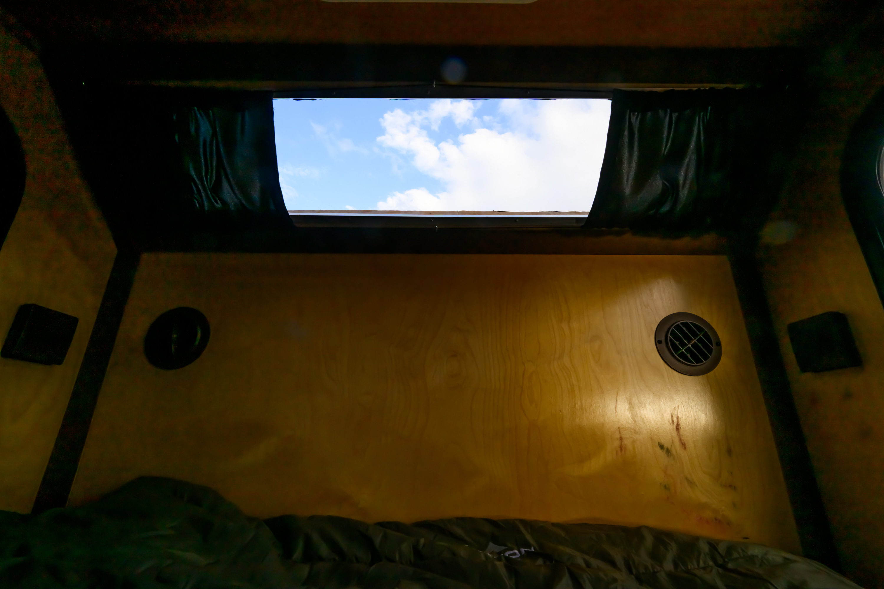 Inside an RV rental: a cozy wooden interior with a window revealing a blue sky and clouds, a bed, and wall vents.. Venture XTR 2022