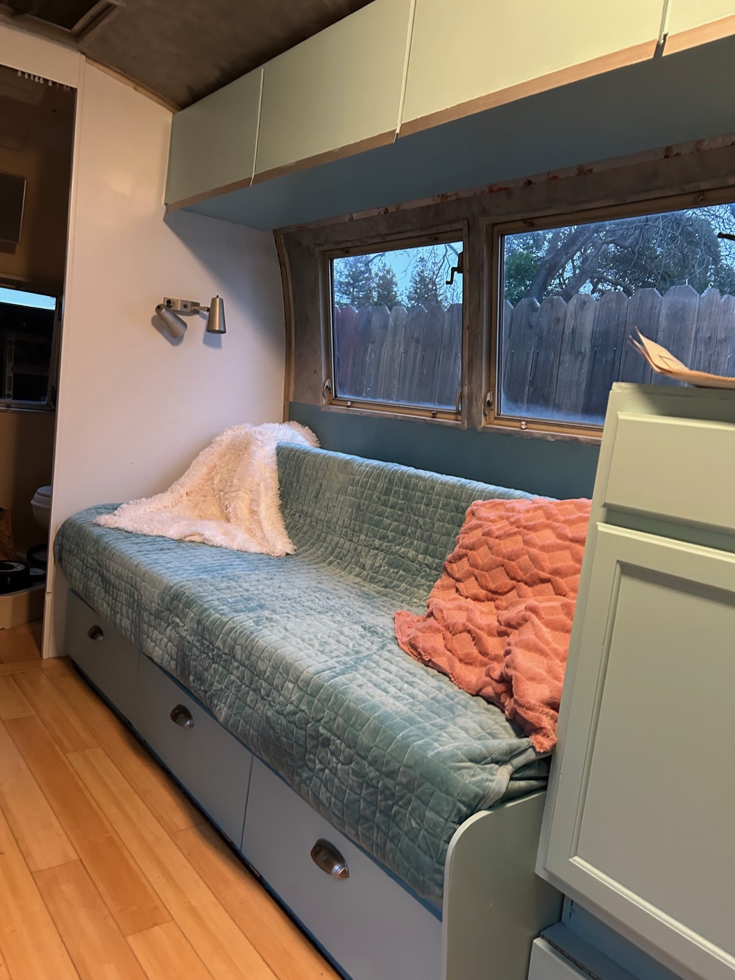 A cozy RV rental interior, featuring a teal daybed with under-bed storage, wall light, and windows viewing a fence.. Airstream Globetrotter 1965