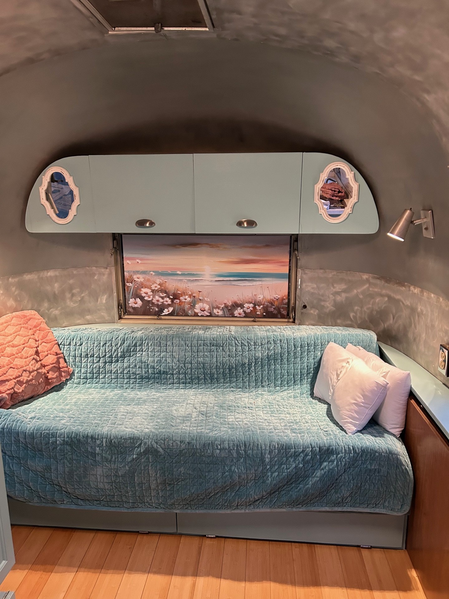 Cozy RV rental interior: light blue sofa bed with pink and white pillows, overhead cabinets, and a beach scene window.. Airstream Globetrotter 1965