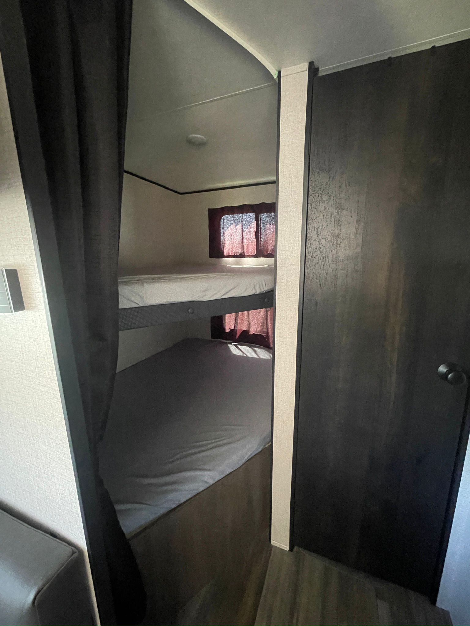 RV rental bunk bed area: stacked beds with light bedding, red-curtained windows, and a dark privacy curtain. Dark door on right.. Starcraft Autumn Ridge 2023
