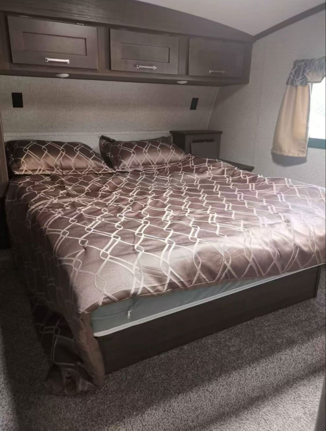 RV rental bedroom showing a bed with patterned brown bedding, overhead storage, a small nightstand, and a carpeted floor.. KEYSTONE LAREDO FIFTH WHEEL 2019