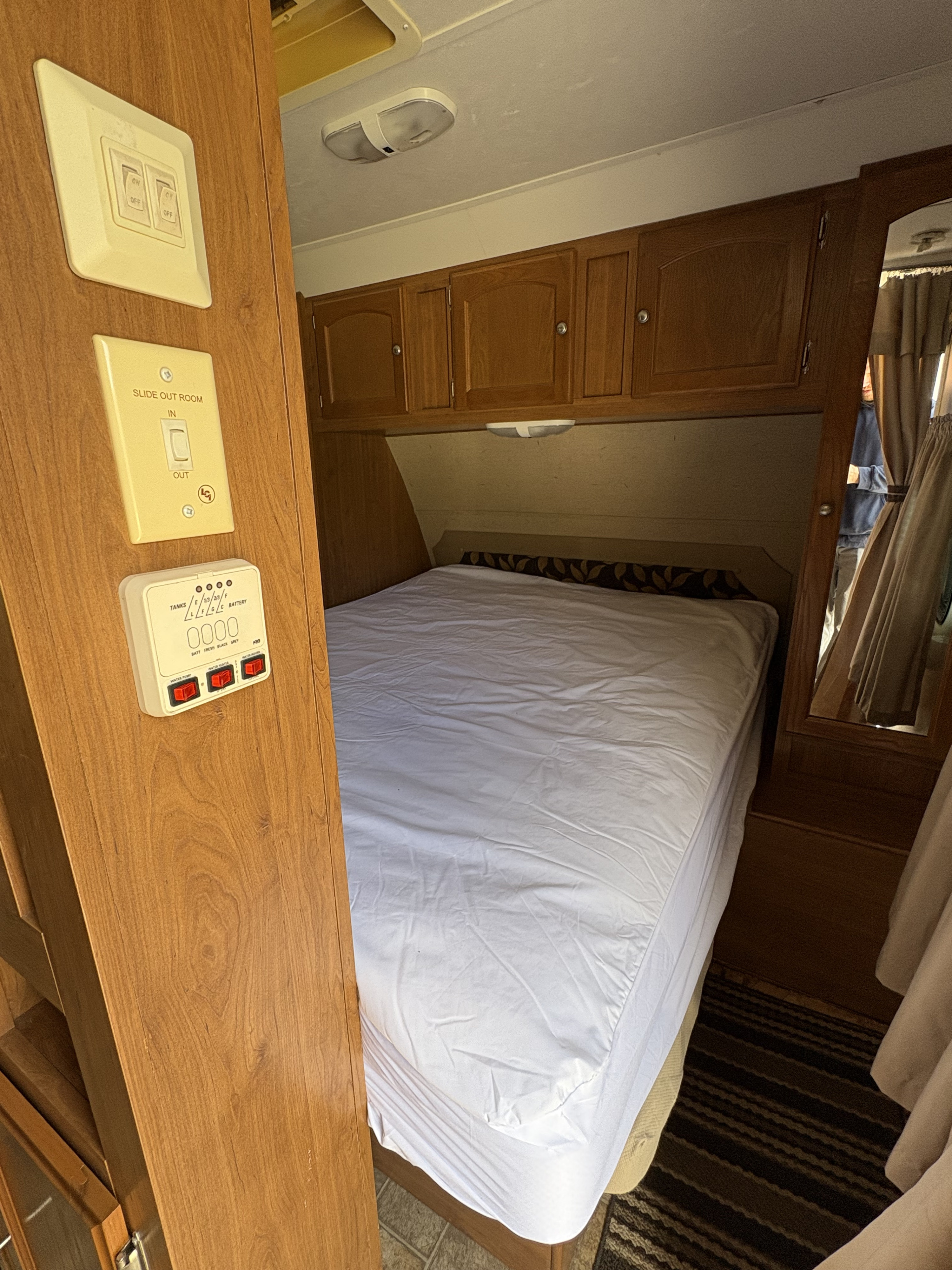 RV rental interior: bedroom with a clean bed, wood cabinets, and a control panel displaying light, slide-out, and tank monitors.. Crossroads RV Sunset Trail 2010