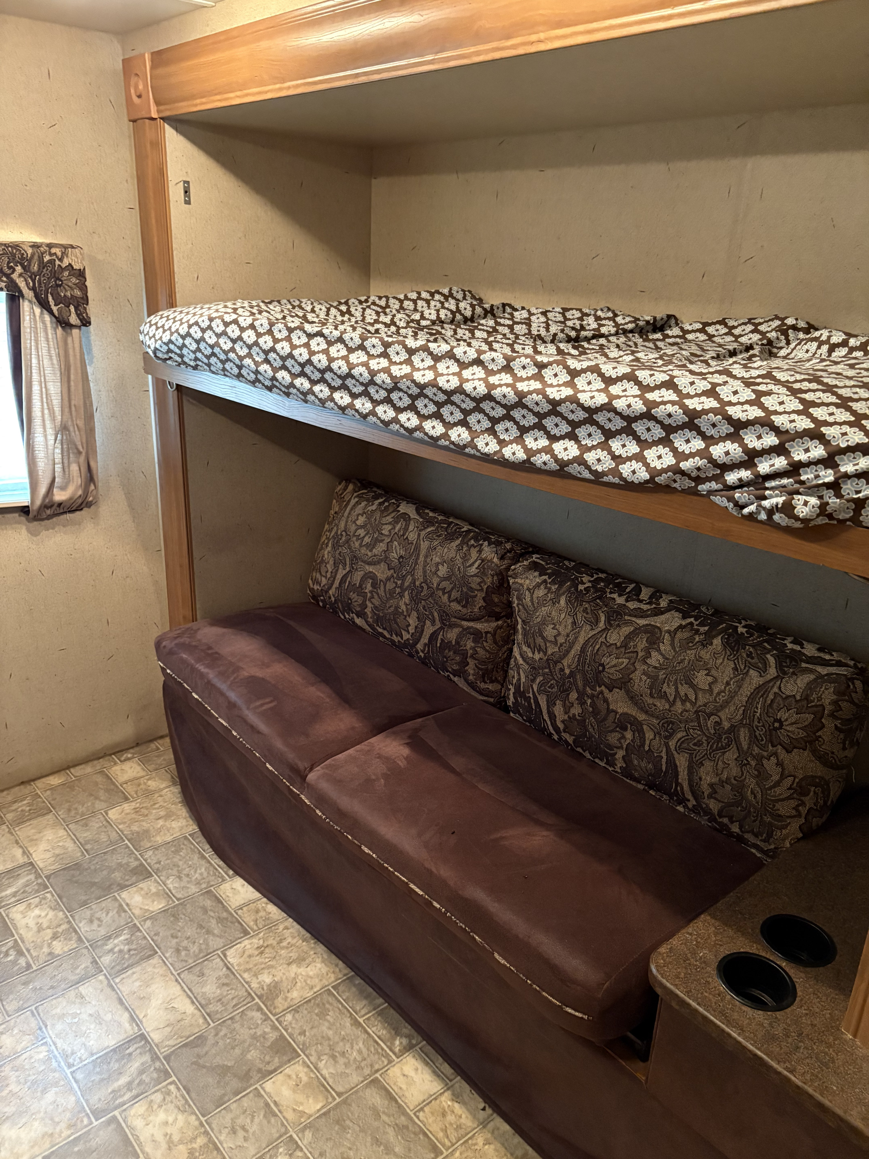 Cozy RV rental: bunk bed with patterned comforter over a brown sofa with pillows and cup holders. Tiled floor.. Crossroads RV Sunset Trail 2010