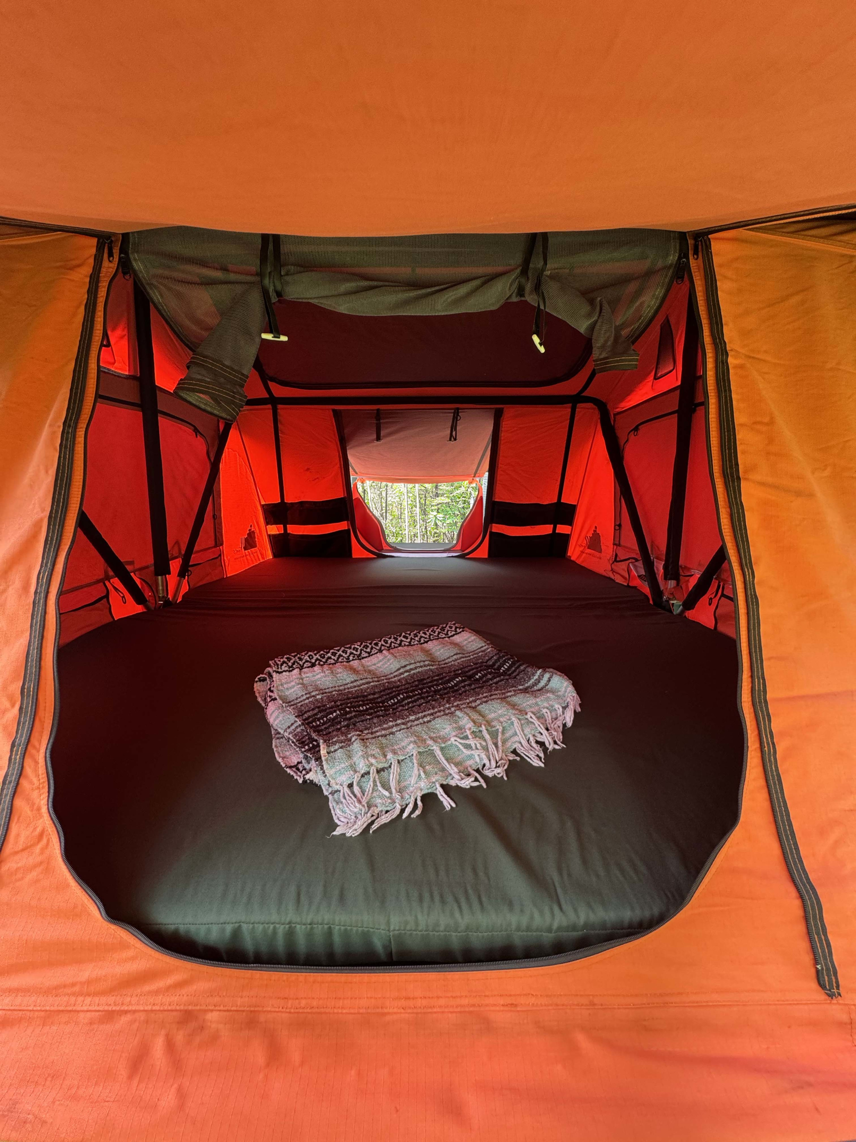 Inside a cozy rooftop tent, perfect for an RV rental adventure. Dark mattress, colorful blanket, view of lush trees.. Borderland Basecamp 2017