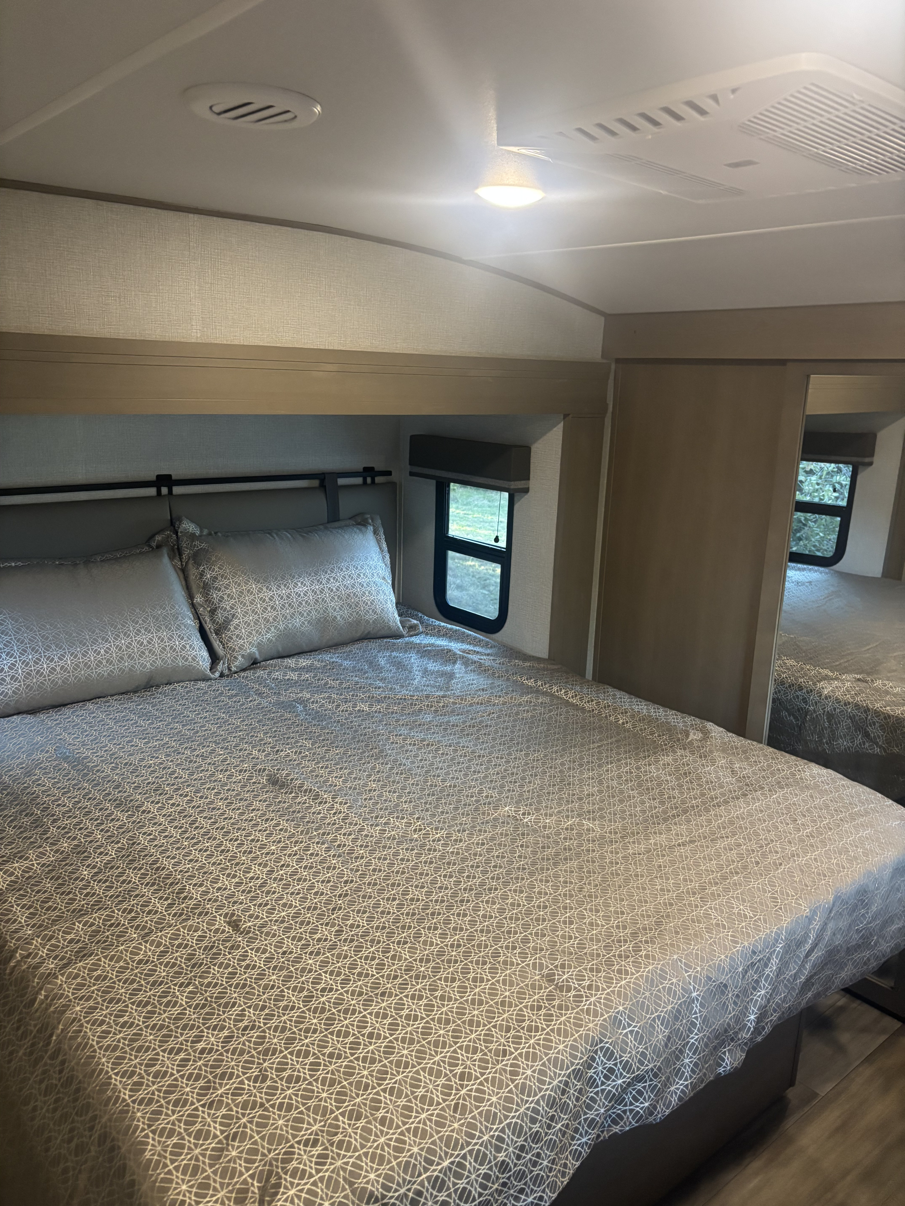 Well-appointed RV rental bedroom. Features include a large bed with gray patterned bedding, compact windows, and ceiling AC.. Grand Design Reflection 2023