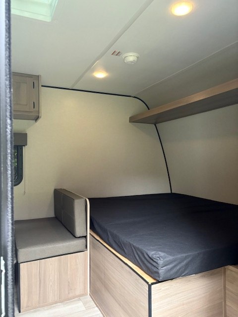 Compact RV rental interior: A bed, bench seating with storage, overhead shelf, and cabinet. Well-lit with a smoke detector.. Keystone RV COLEMAN 17BX 2024