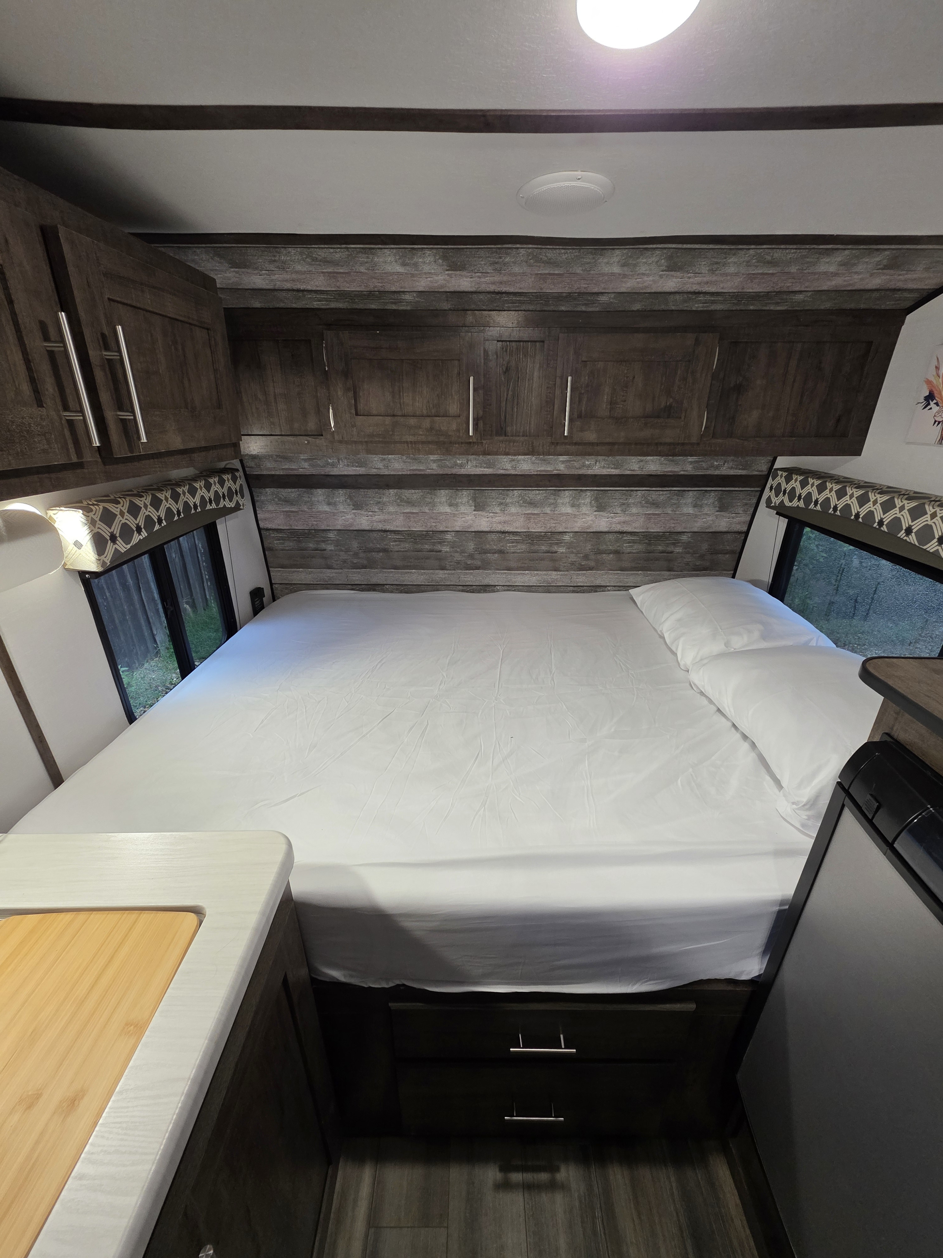 Cozy RV rental bedroom with a neatly made bed, wood cabinets, and windows offering nature views.. Riverside RV Retro 2021