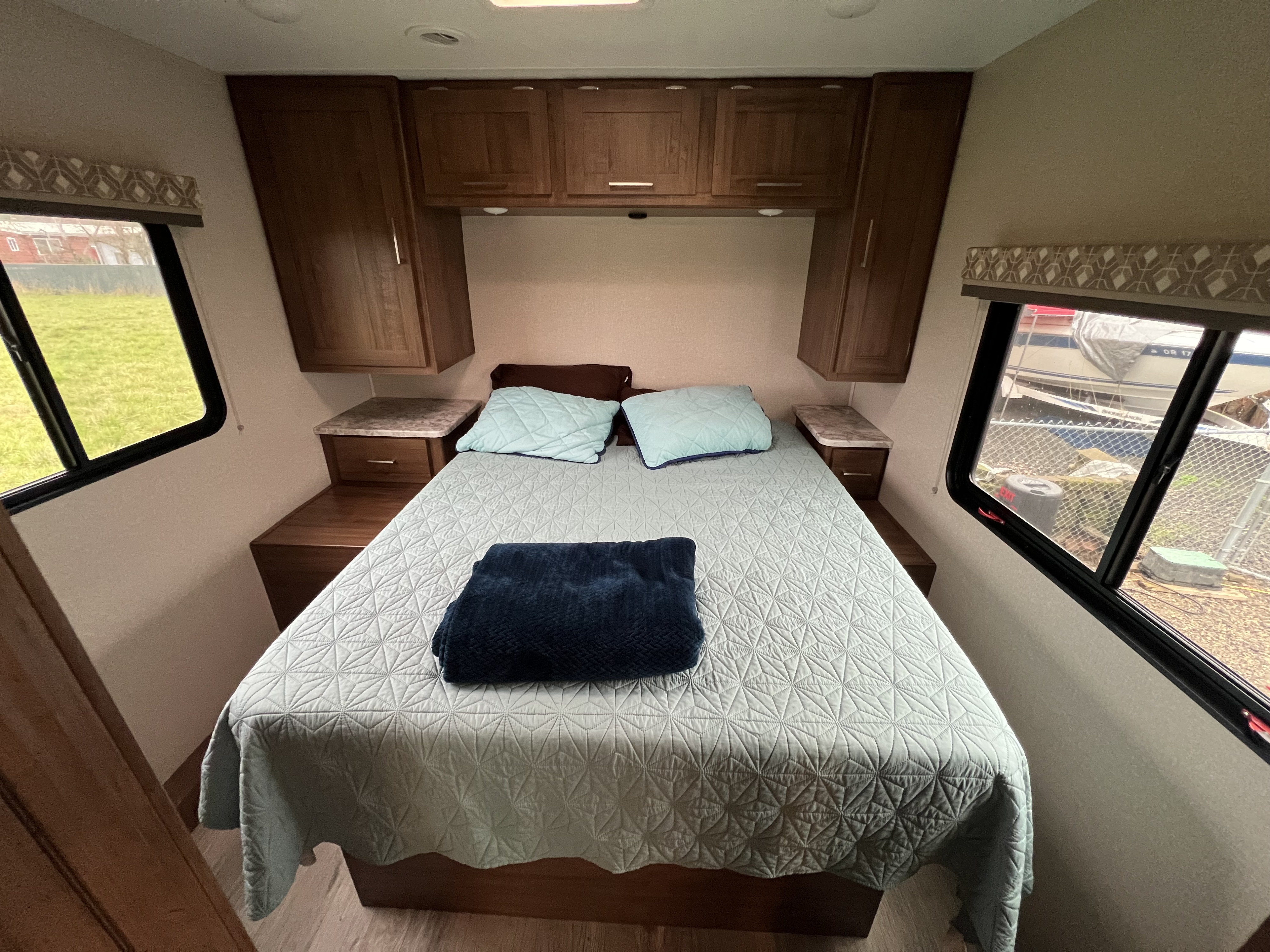 Cozy RV rental bedroom with a light blue quilt on a queen bed, flanked by wooden cabinets and large windows.. Coachman RV Pursuit 27 XPS 2020