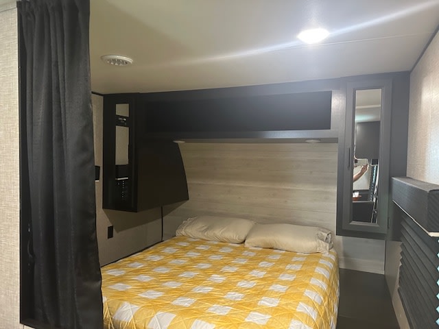 Compact RV rental bedroom featuring a bed with yellow quilt, dark curtains, and smart overhead storage. Ideal for travelers.. Jayco Jay Flight 2023
