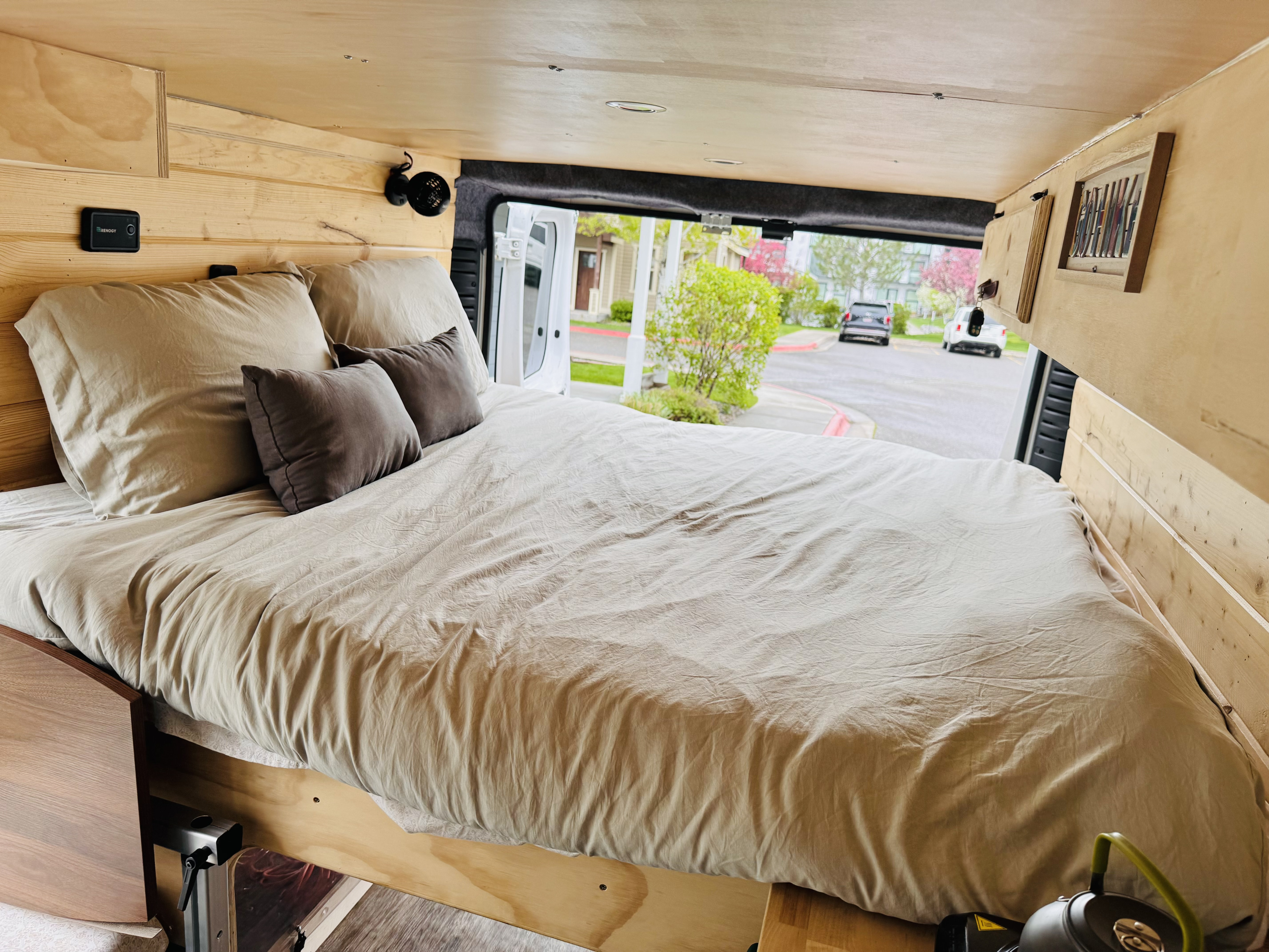 Cozy RV rental interior: a comfy bed with light brown sheets, wooden walls, and a view out the back doors.. RAM Promaster 2017